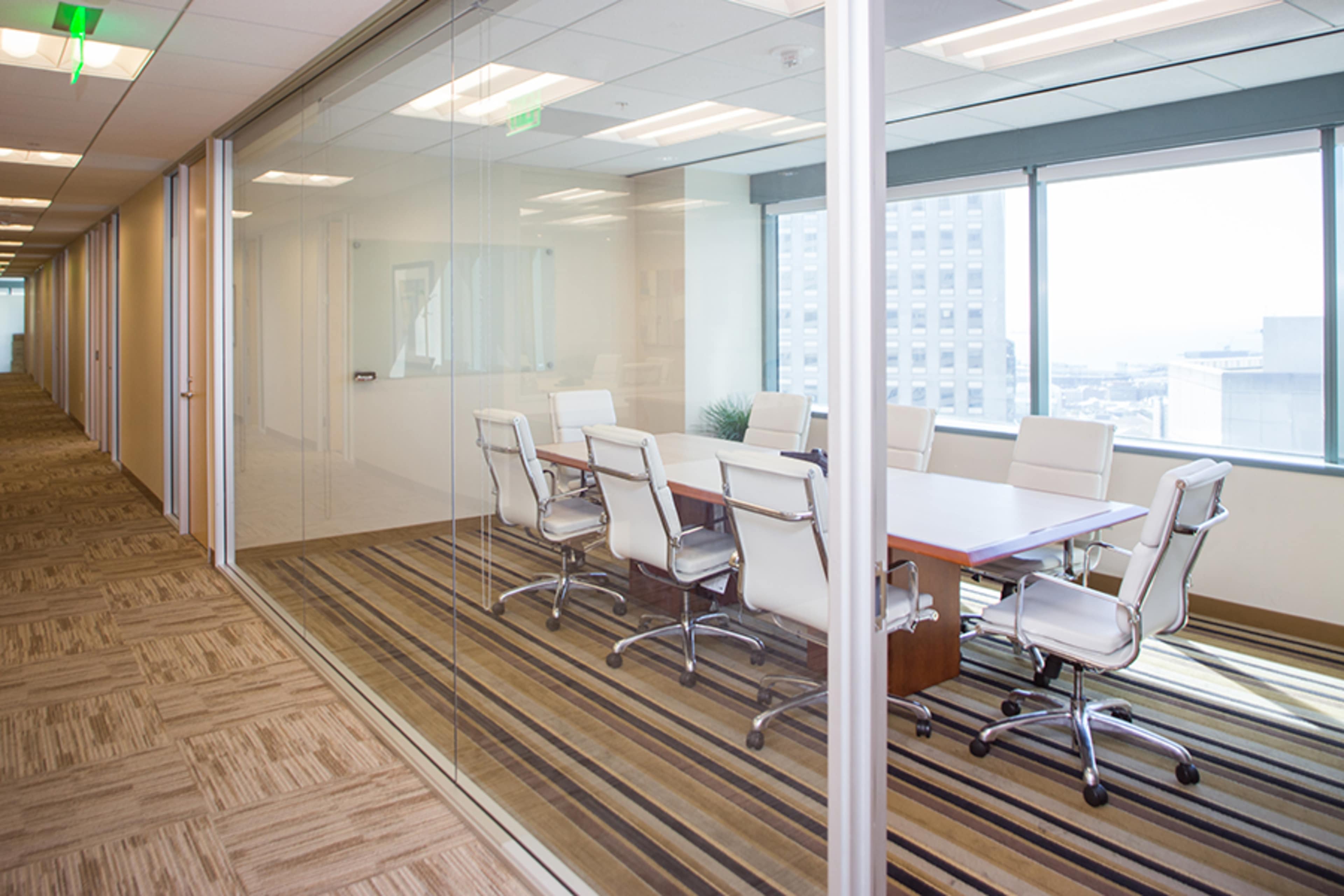 A glass-walled conference room with a large table and white chairs is visible from a carpeted corridor.