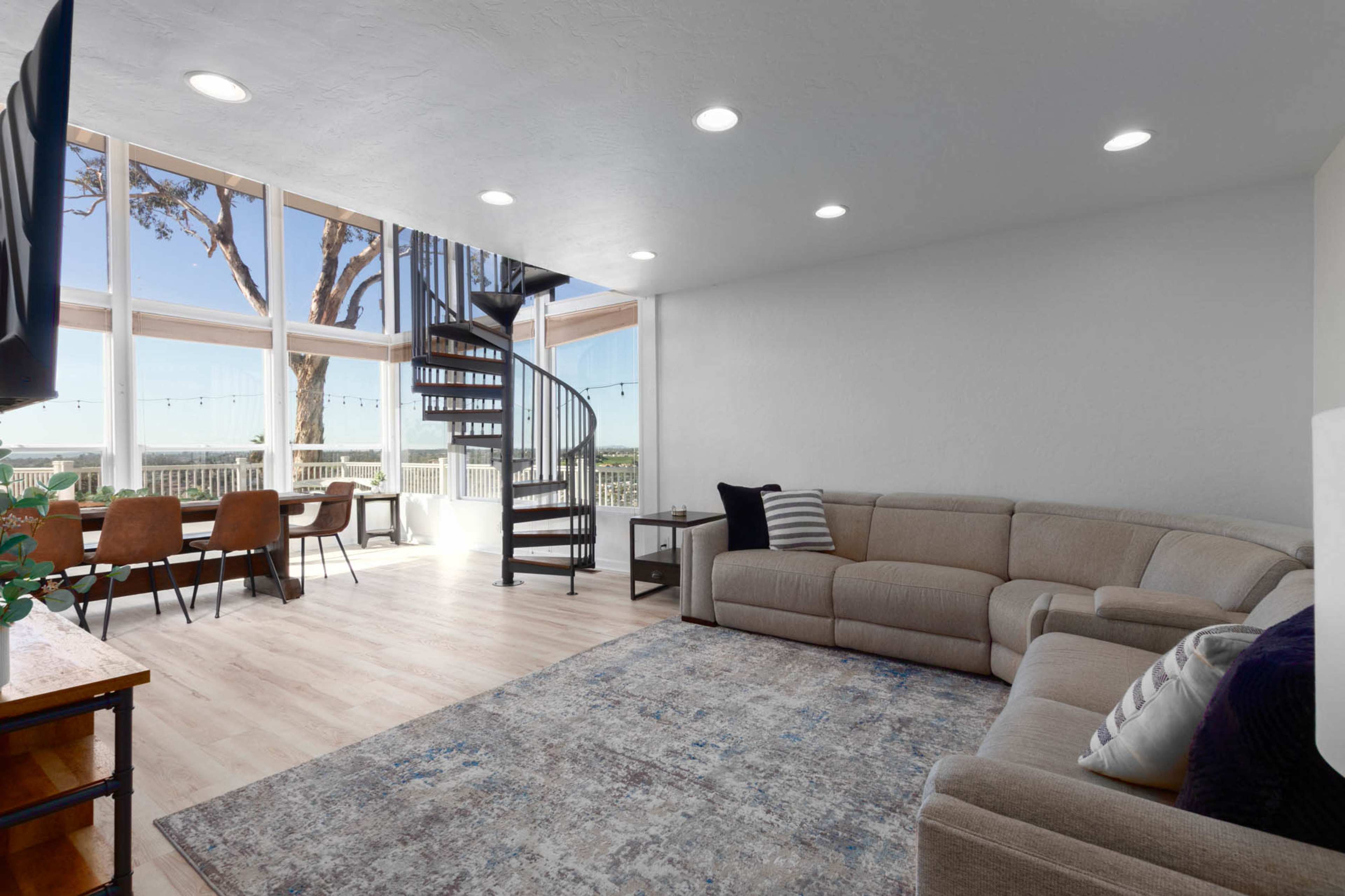 The image shows a modern living room with a spiral staircase, a sectional sofa, and a dining area featuring a view through large windows.
