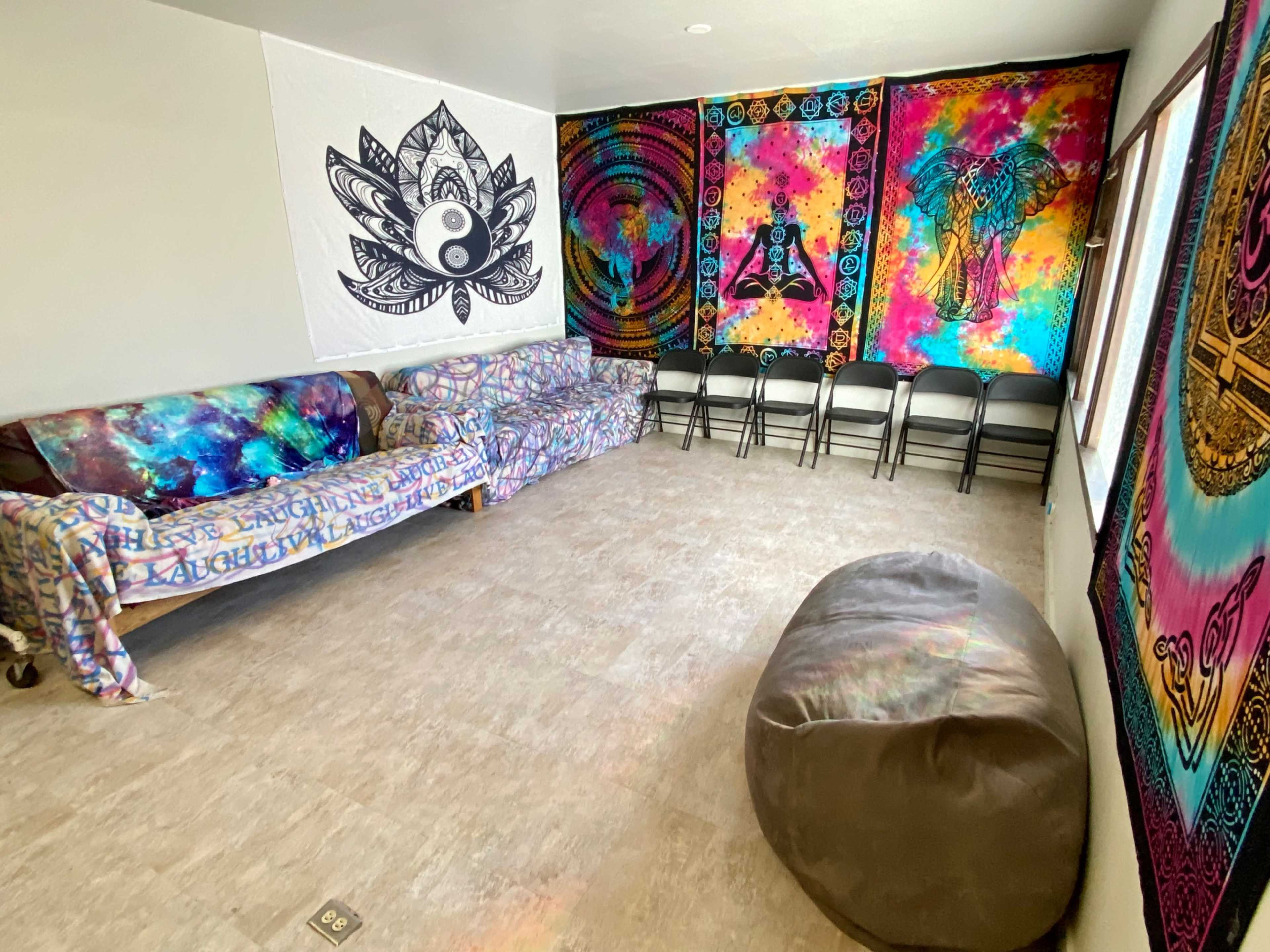 A brightly colored room with patterned wall tapestries, a sofa covered in a printed throw, several folding chairs, and a large bean bag on the floor.