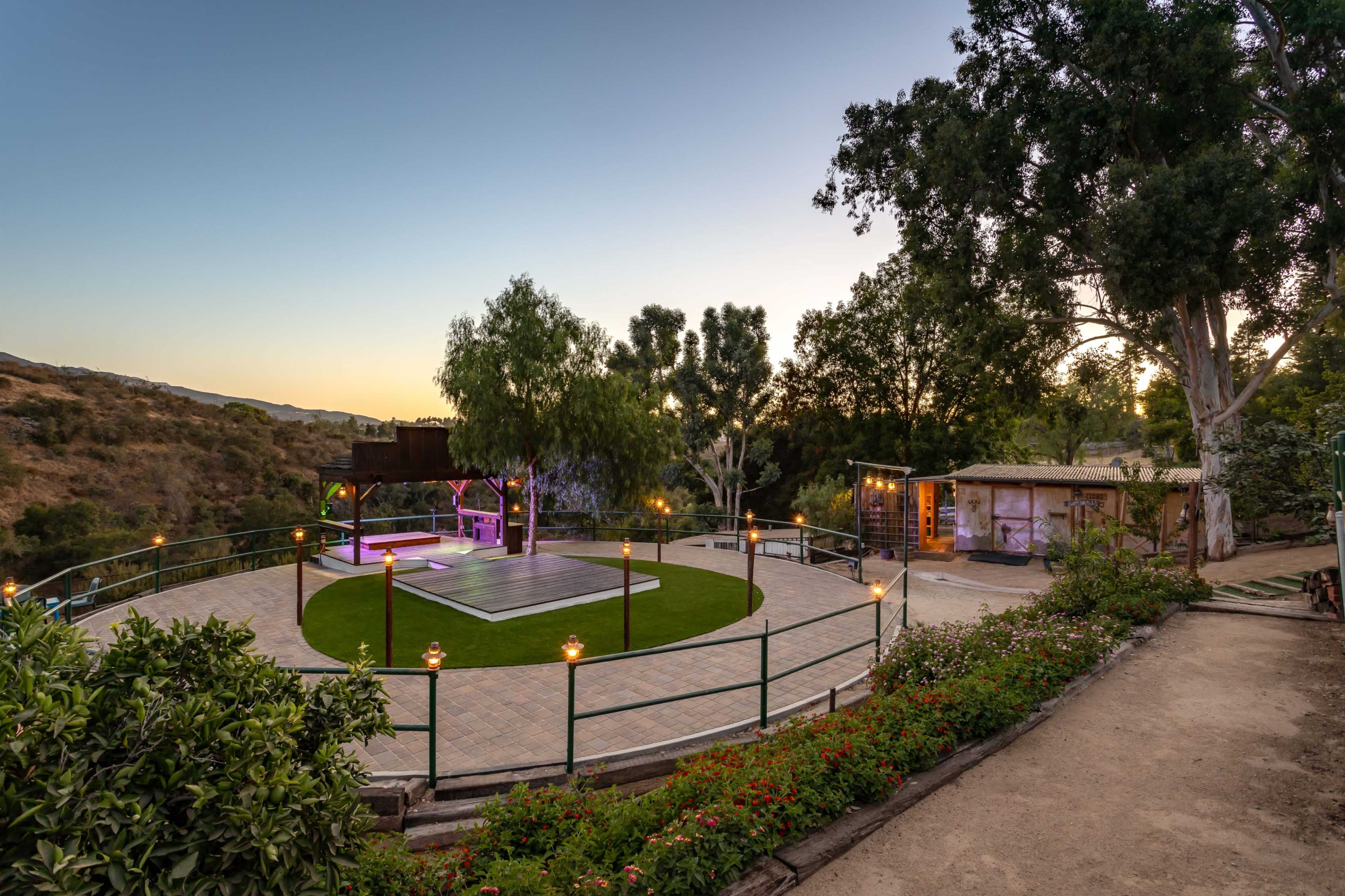 A landscaped outdoor venue with a circular stage, surrounded by greenery and a building in the background, set against a twilight sky.