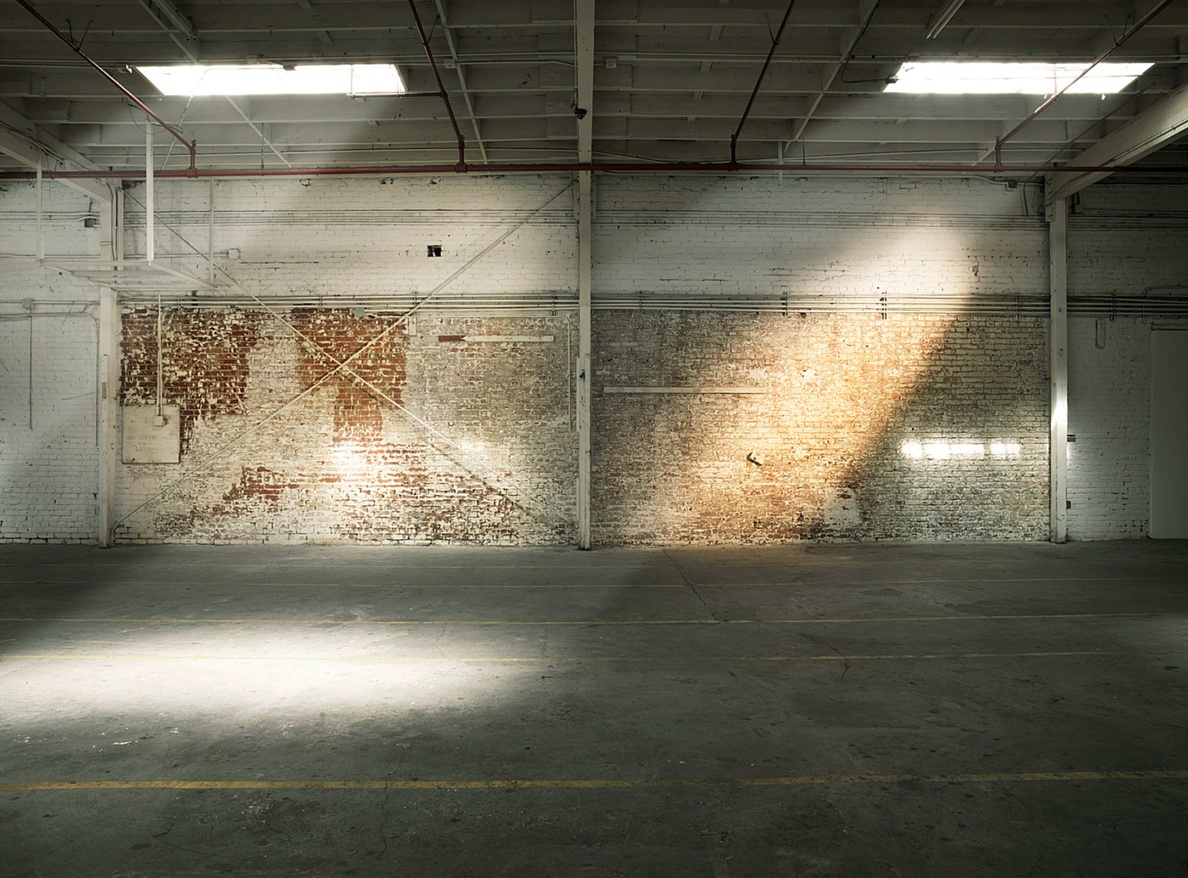 The image shows a large, empty warehouse with a textured brick wall and sunlight streaming through a window, casting a beam of light on the floor.