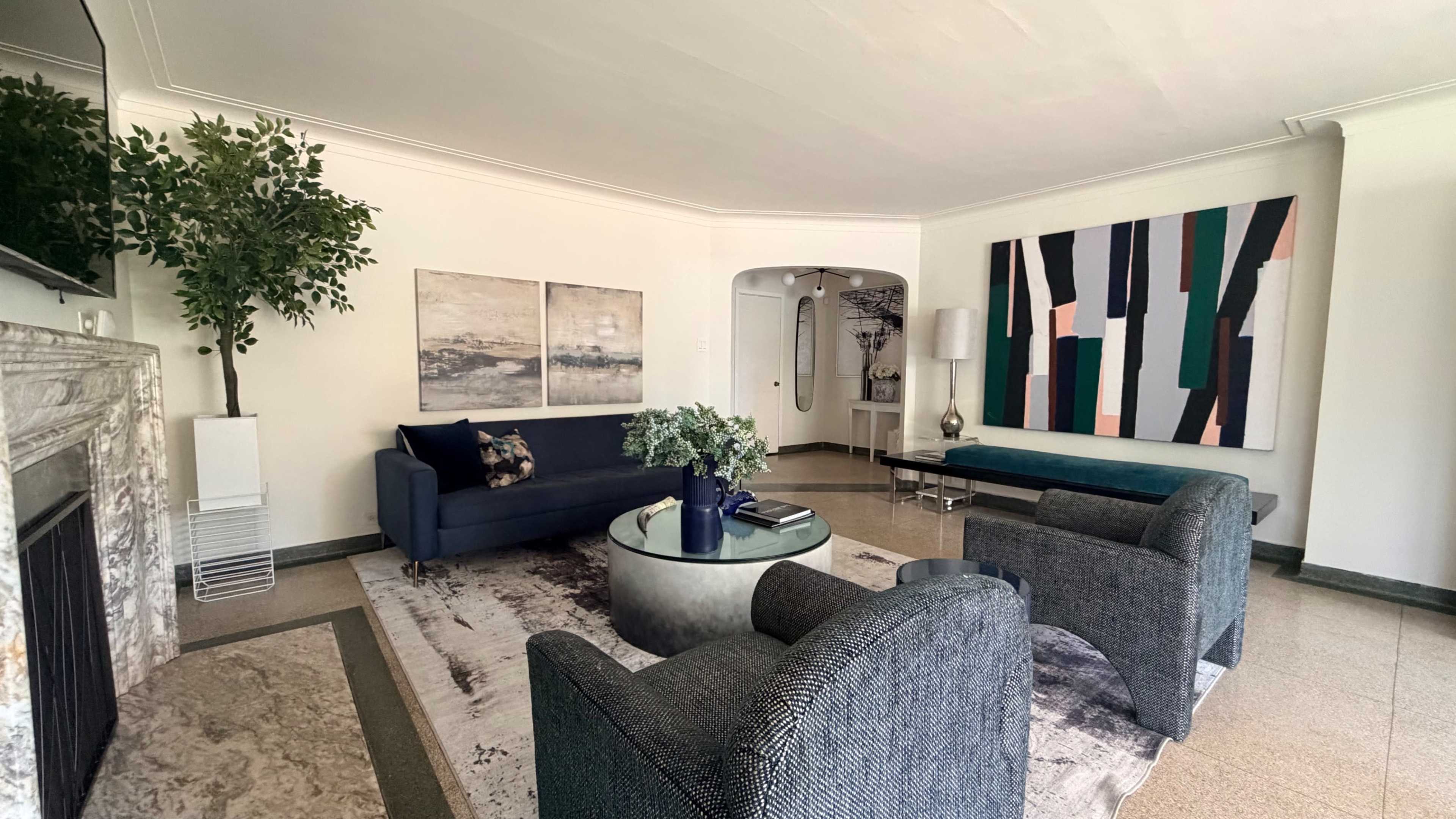 The image shows a modern living room featuring a dark sofa, two patterned armchairs, a circular coffee table, and artwork on the walls, with a large window providing natural light.