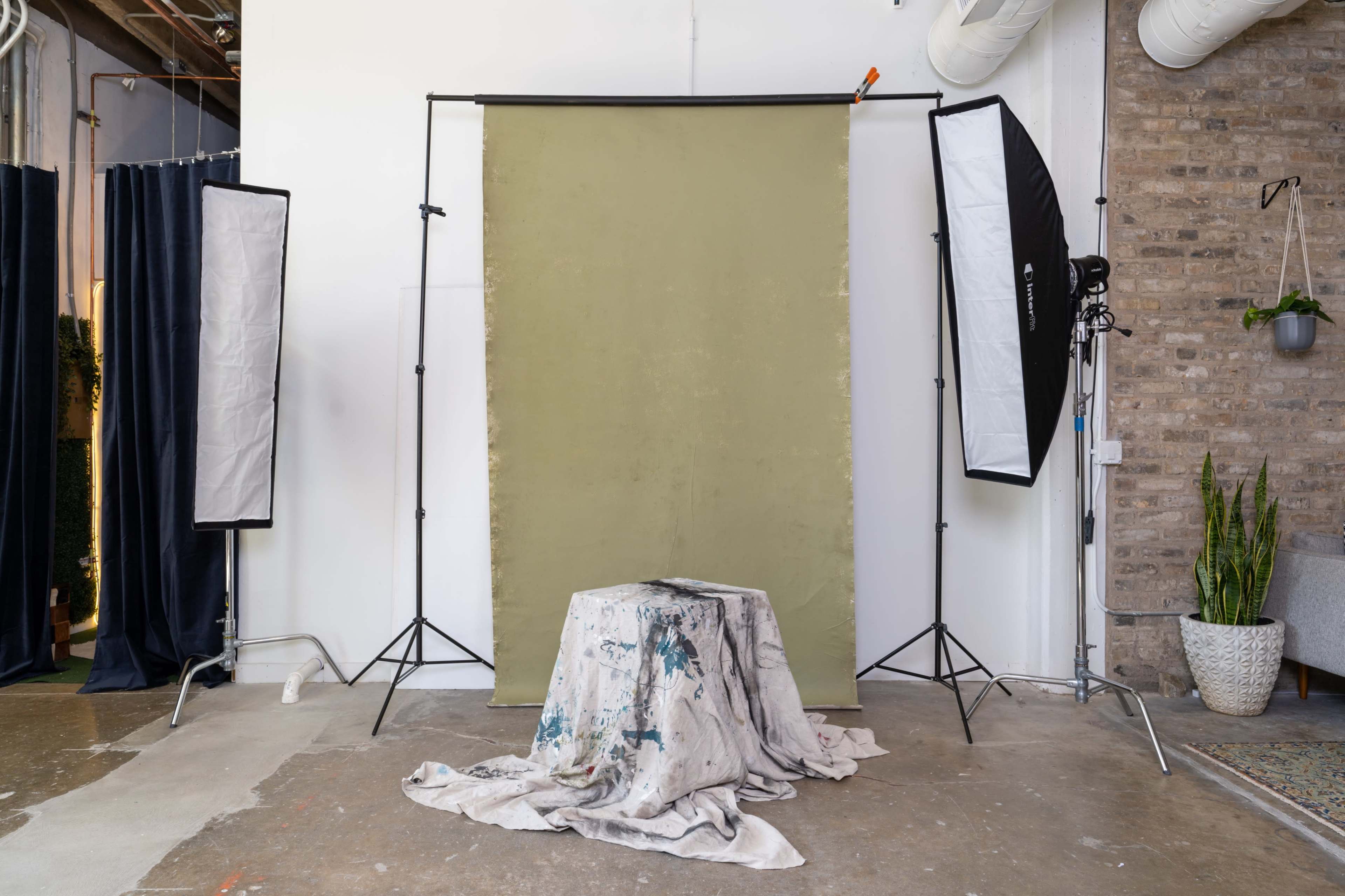 A photography studio is set up with a green backdrop, two softbox lights, and a draped table in the center.