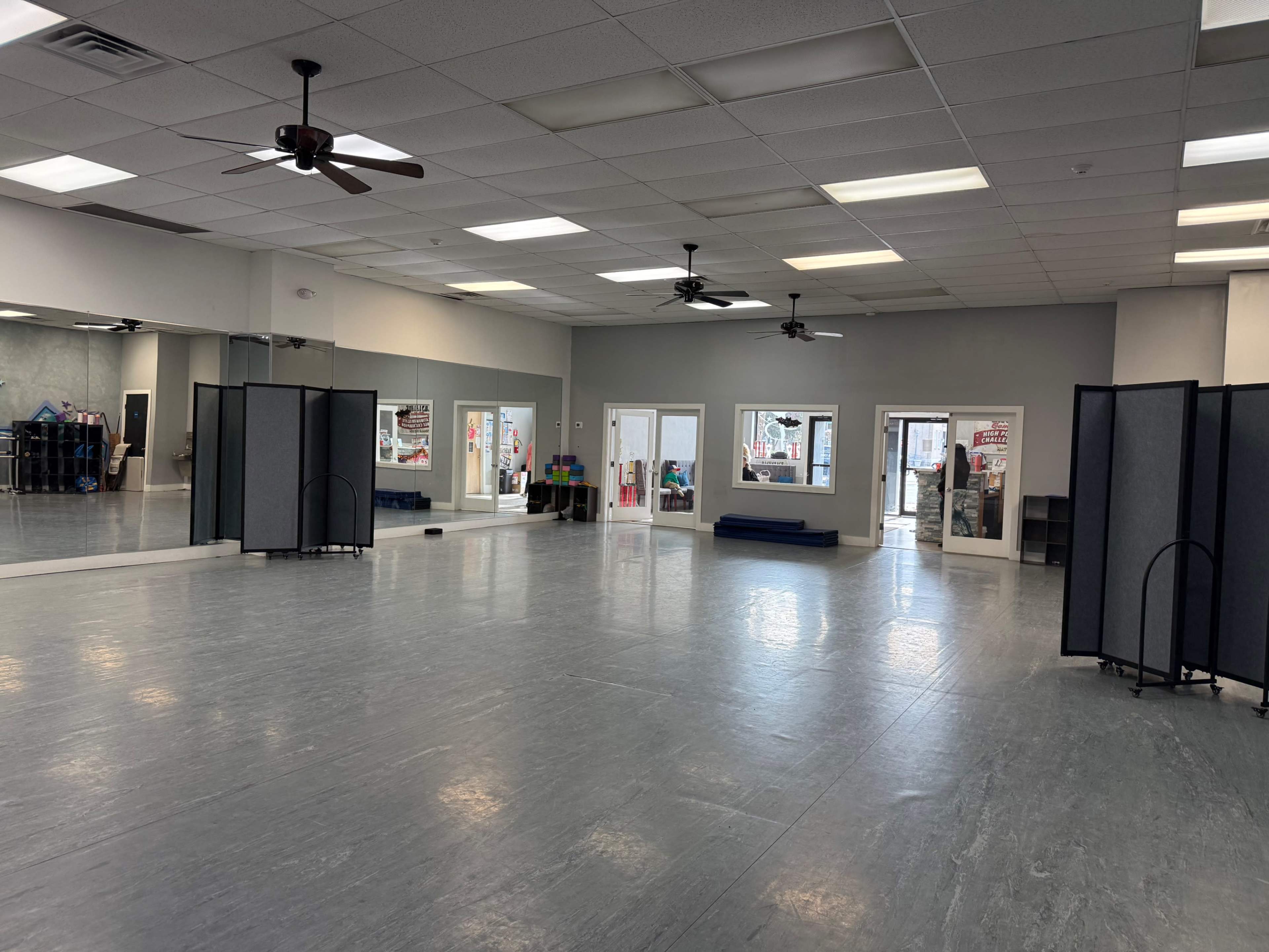 The image shows a spacious, empty room with a polished floor, several ceiling fans, large mirrors on one wall, and movable partition screens.