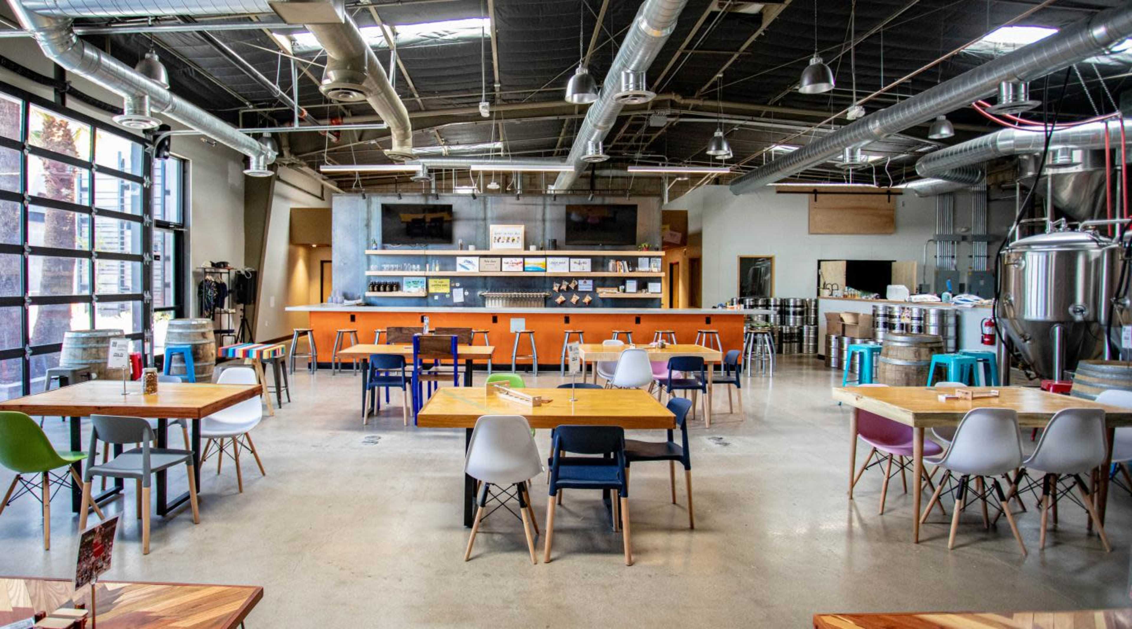 The image shows a spacious, modern dining area with multiple tables and chairs arranged around a central bar and brewing equipment in the background.