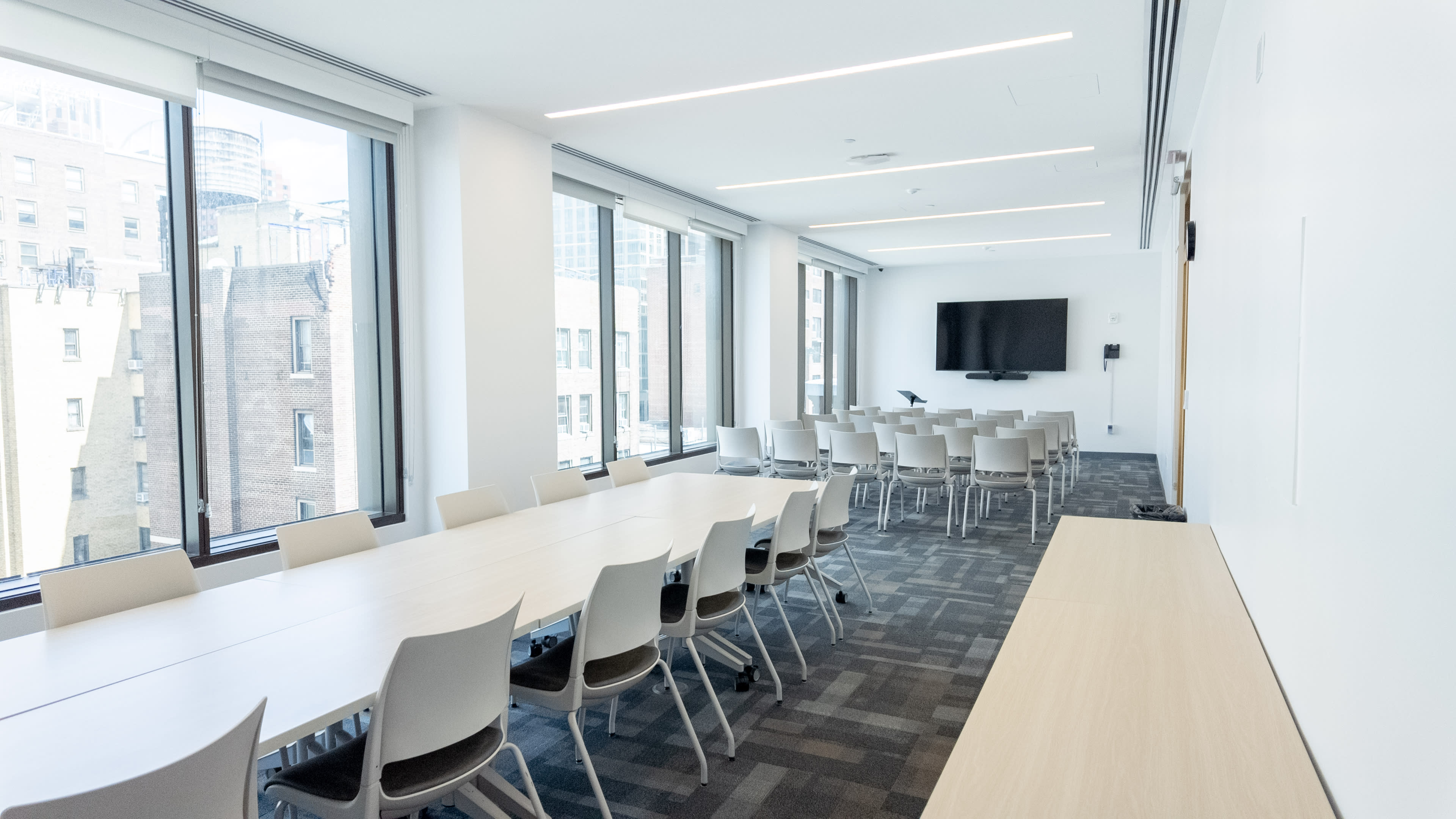 The image shows a modern meeting room with a long wooden table, multiple chairs, large windows, and a wall-mounted television.