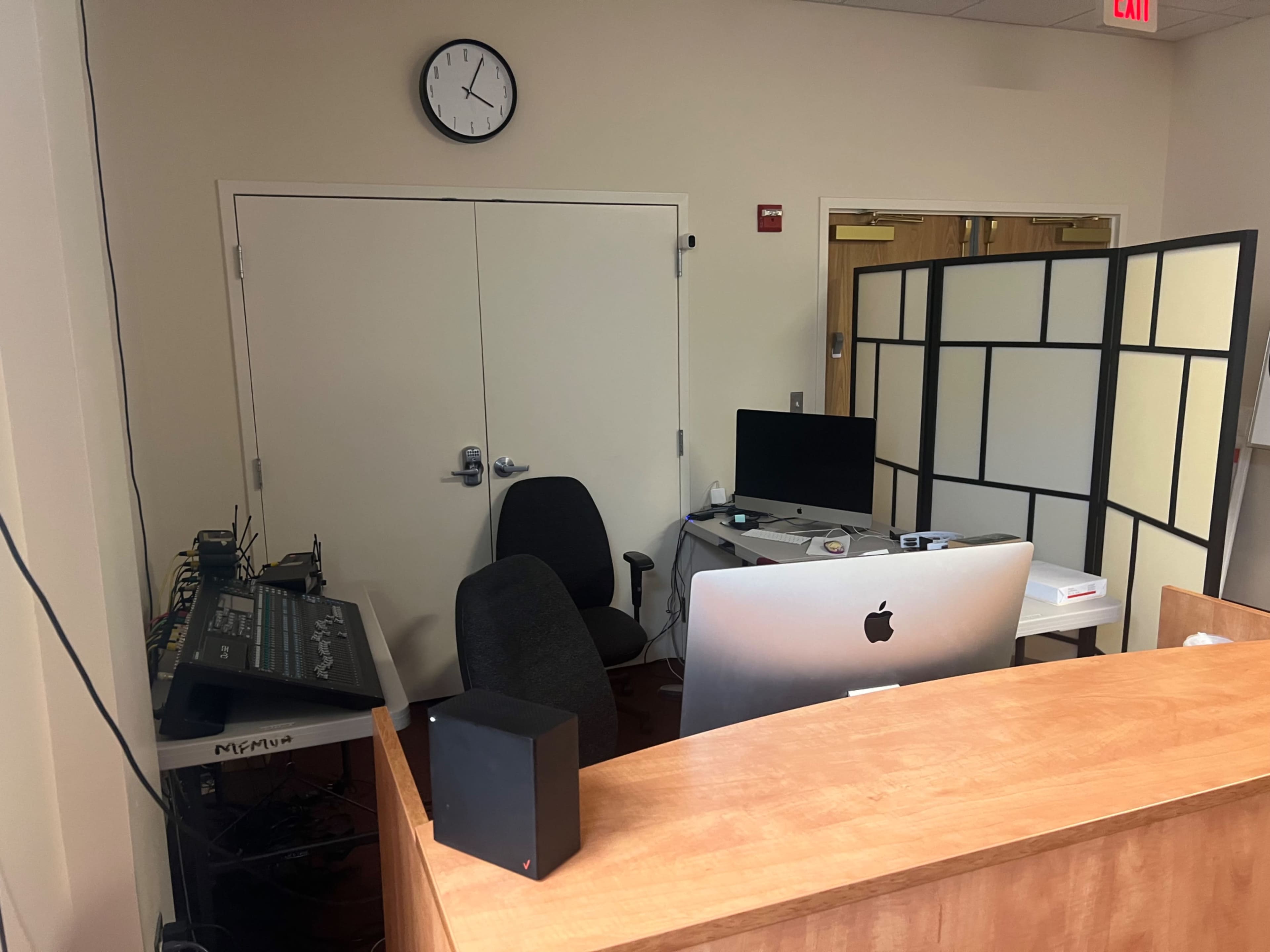 The image shows an office space with a wooden reception desk, a computer setup, a sound mixing board, and a partition screen.