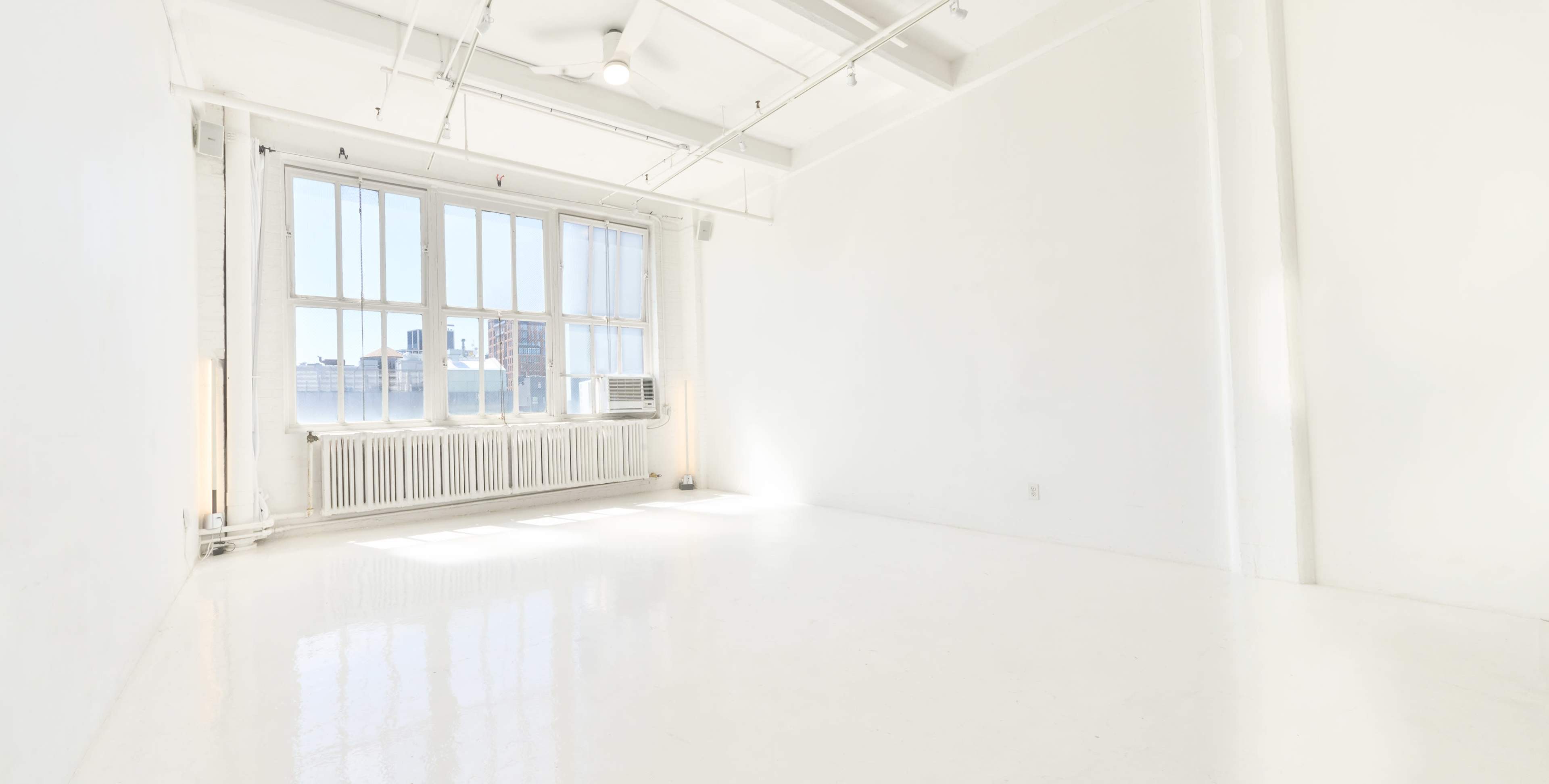 The image shows a bright, empty room with white walls and a polished floor, featuring large windows along one side.
