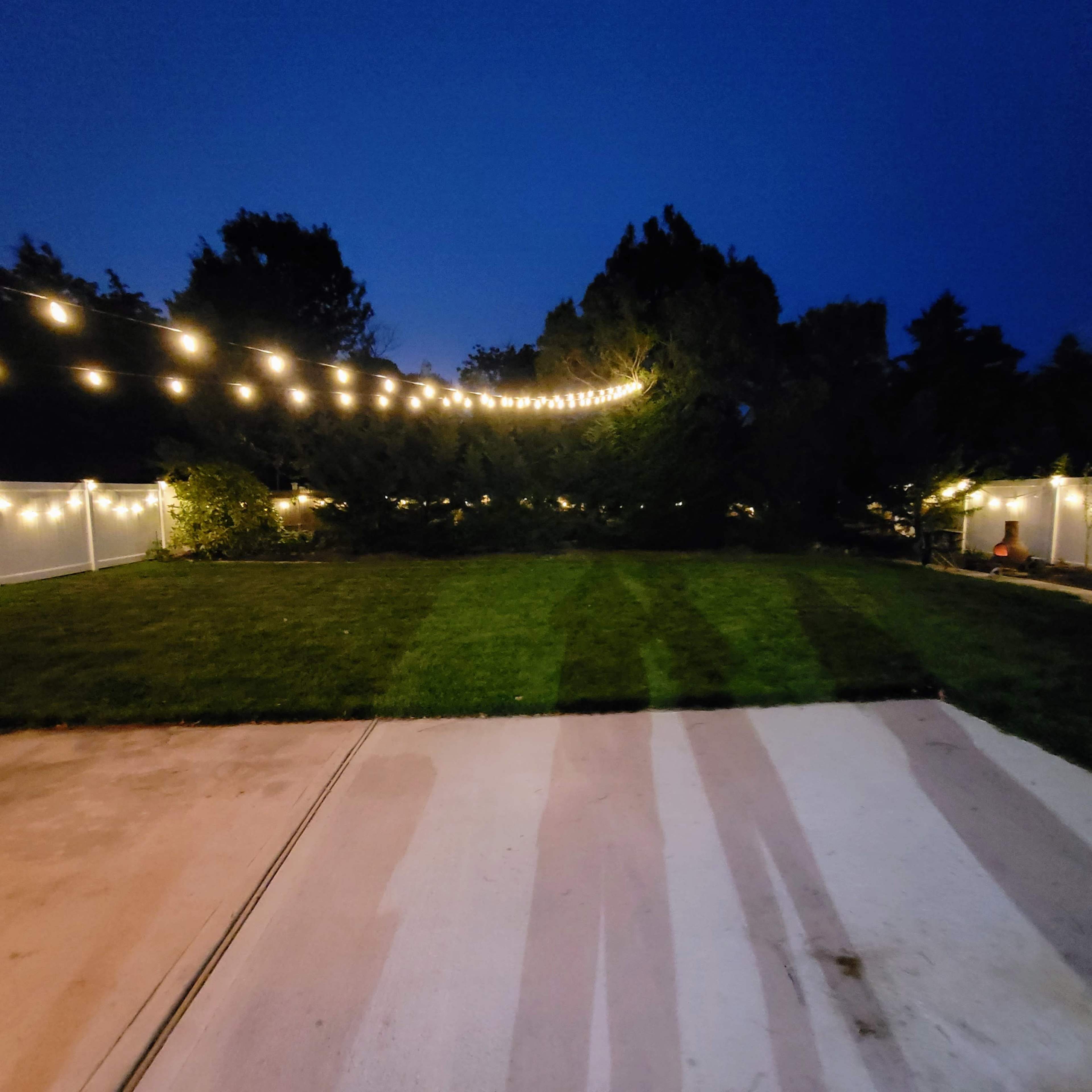 A well-lit backyard features string lights hung over a grassy area with a concrete patio in the foreground.