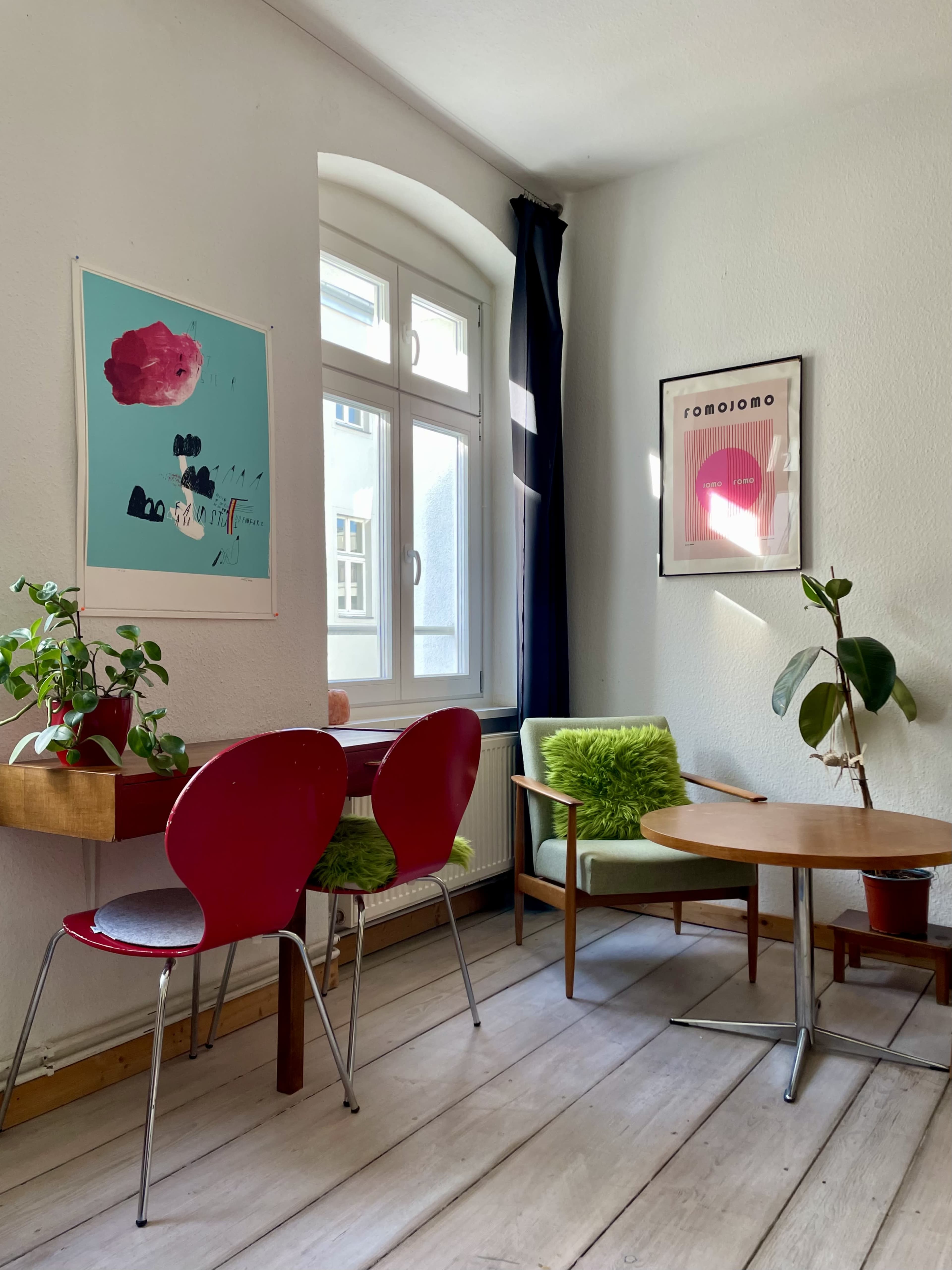 The image shows a cozy interior featuring a small dining table, colorful chairs, and framed artwork on the walls.