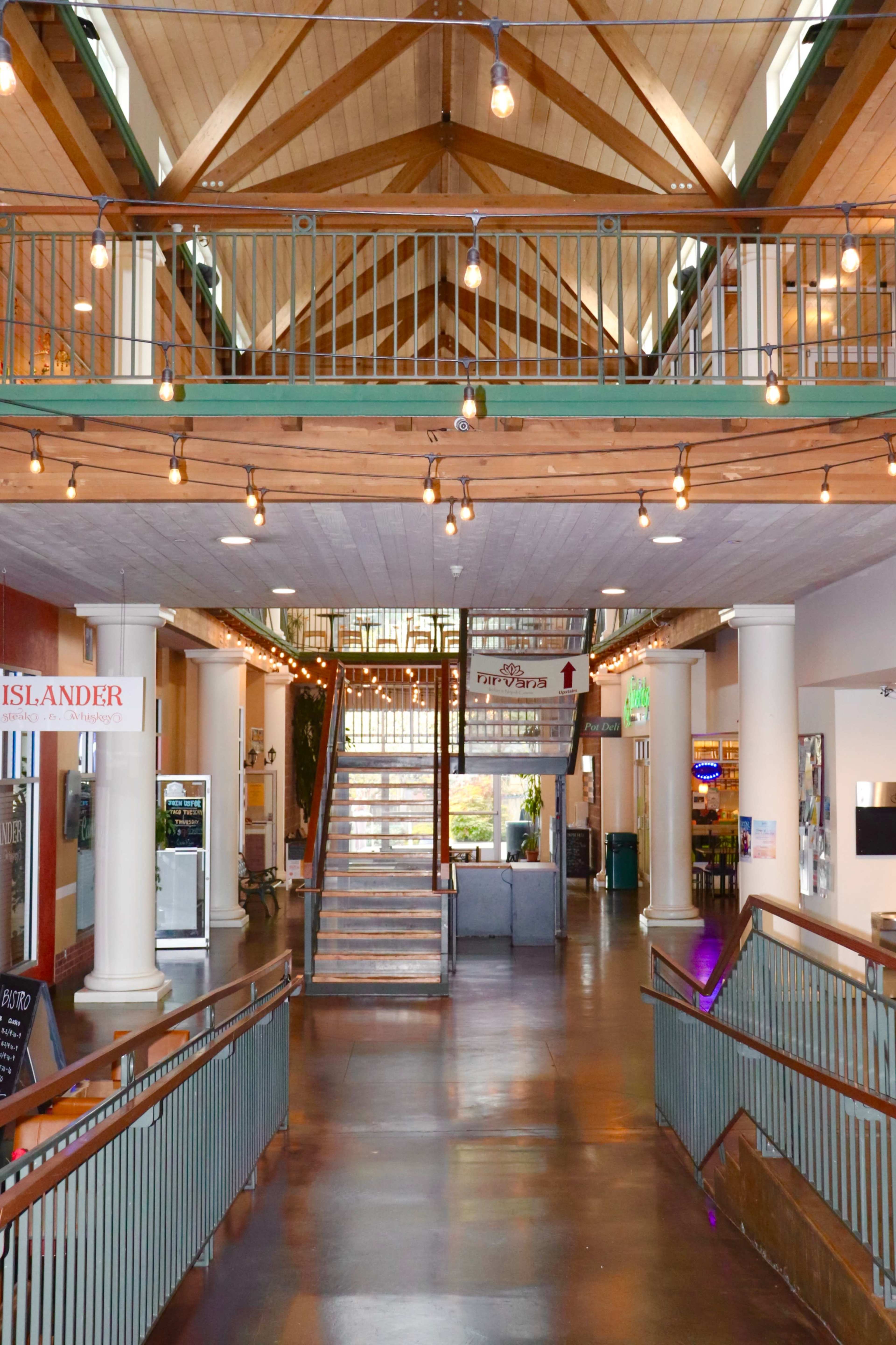 The image shows a spacious indoor area with a high wooden ceiling, a staircase in the center, and multiple businesses lining the walls.