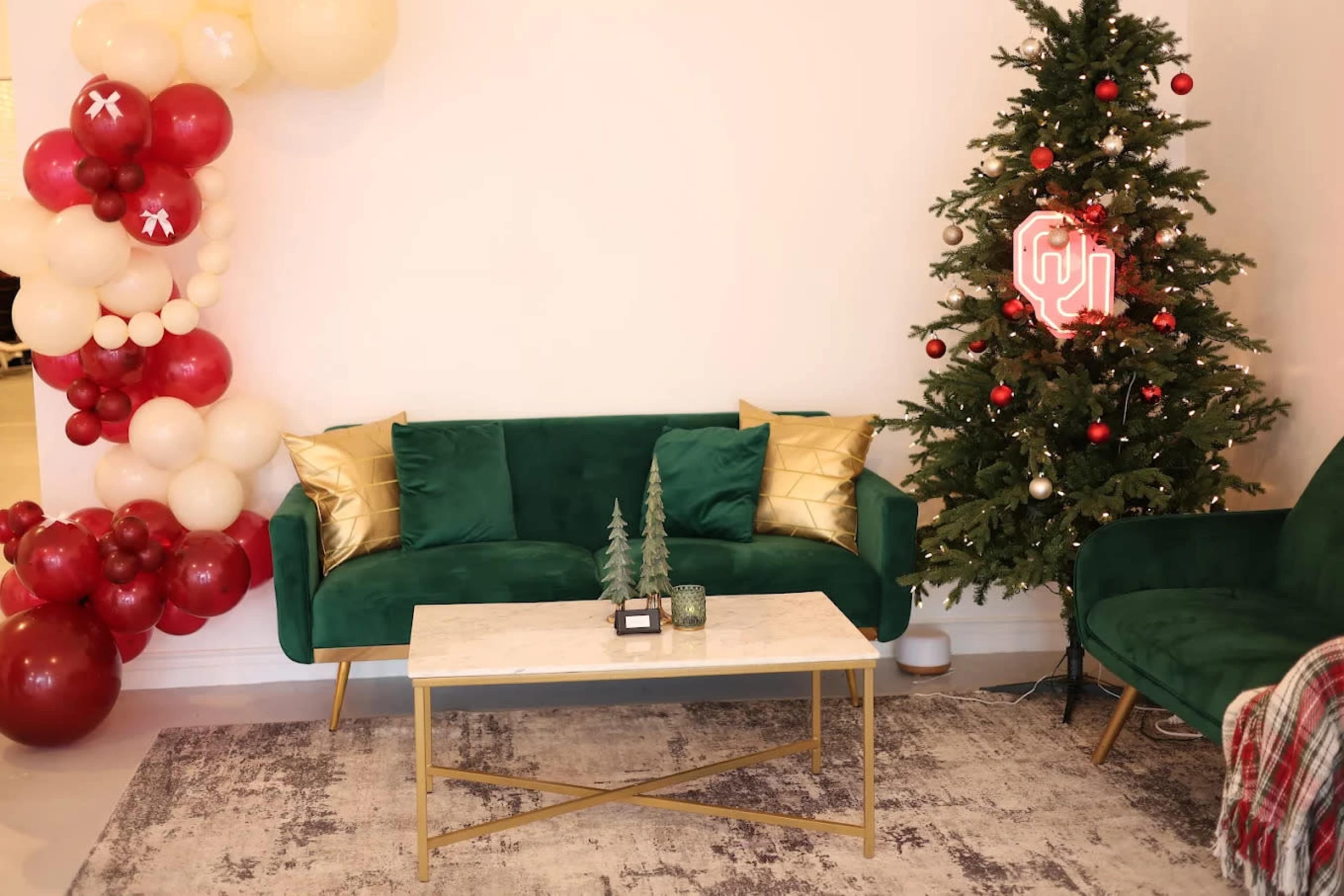 A cozy living room features a green velvet sofa, a marble coffee table, decorated Christmas tree, and festive balloon garland.
