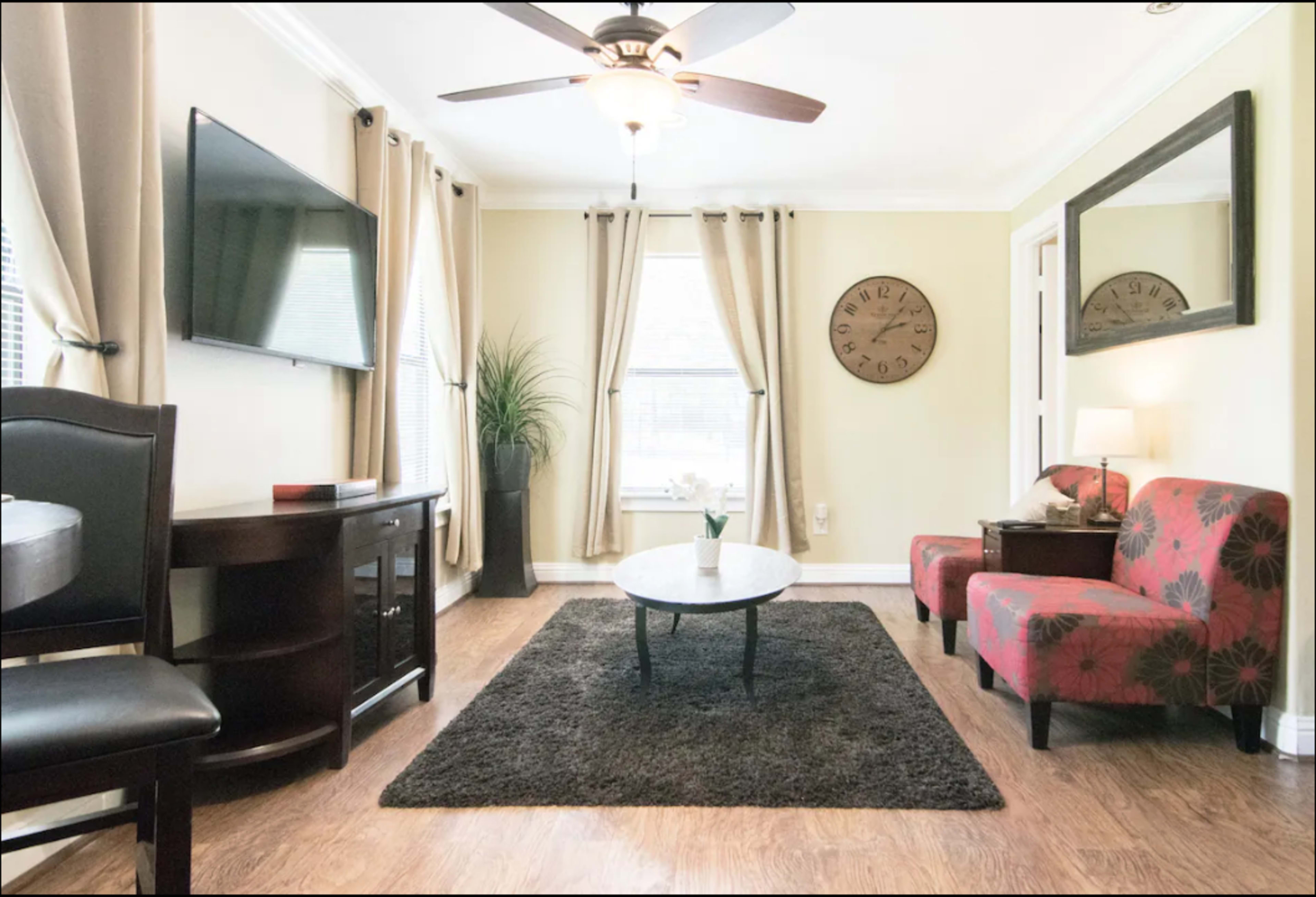 A living room features a television mounted on the wall, a round coffee table on a dark rug, and a pair of patterned chairs flanking a side table.