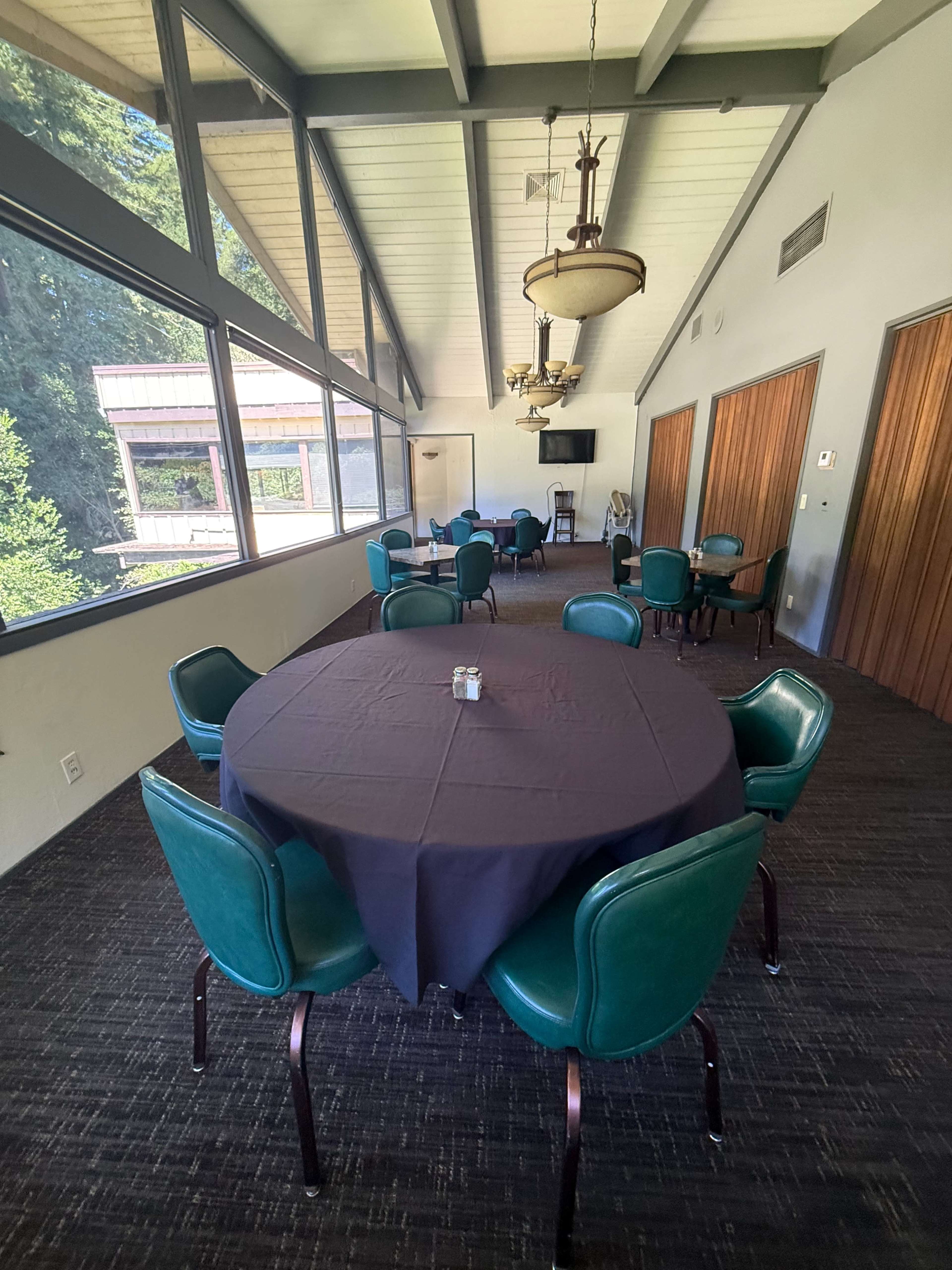 A meeting room with a black tablecloth-covered round table surrounded by green chairs, featuring large windows that provide a view of trees outside.