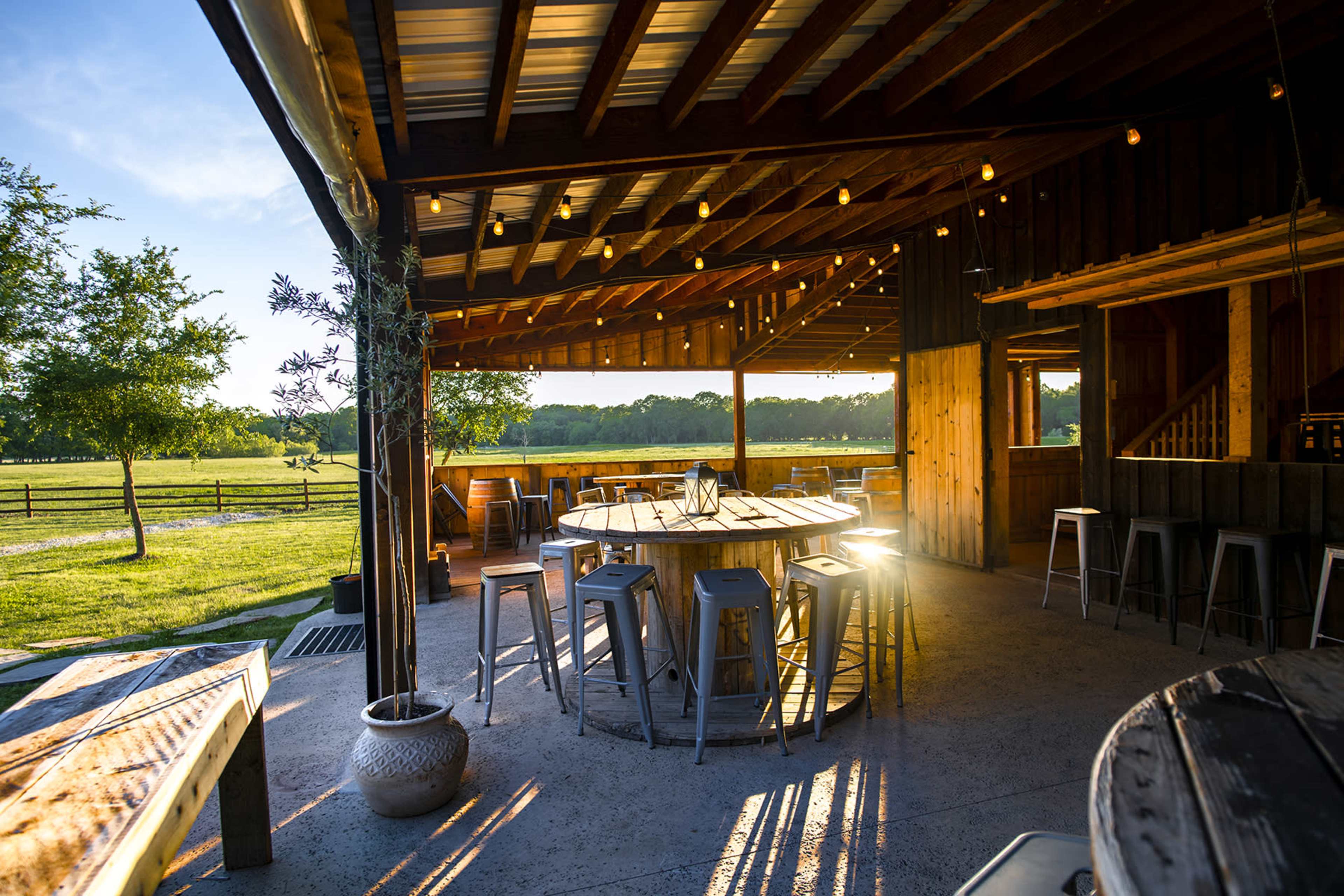 A rustic outdoor seating area features a large round table surrounded by metal stools, illuminated by warm sunlight, with a scenic field visible in the background.