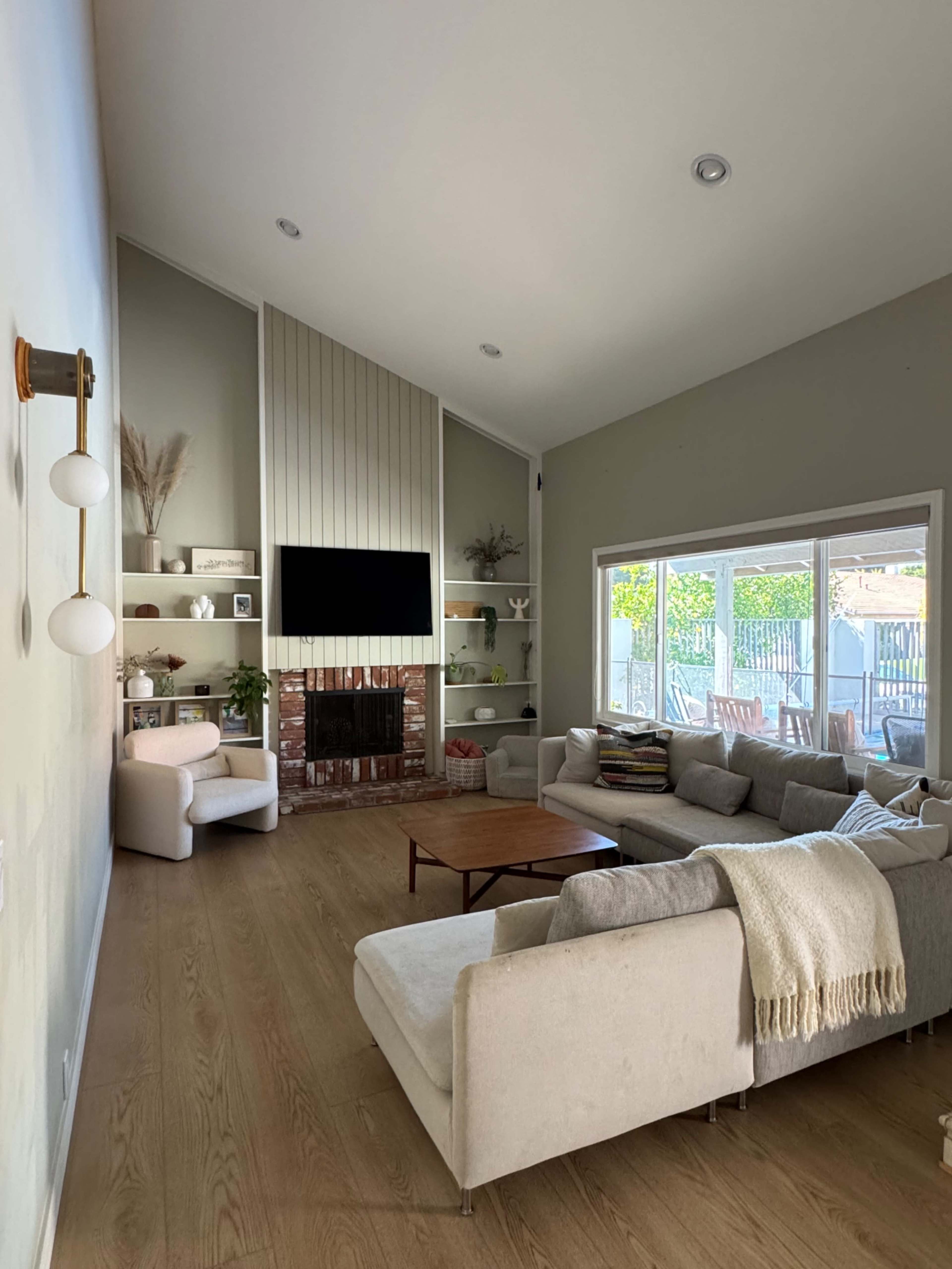A spacious living room features a gray sectional sofa, a wooden coffee table, and a large window overlooking a patio, with a television mounted above a brick fireplace.