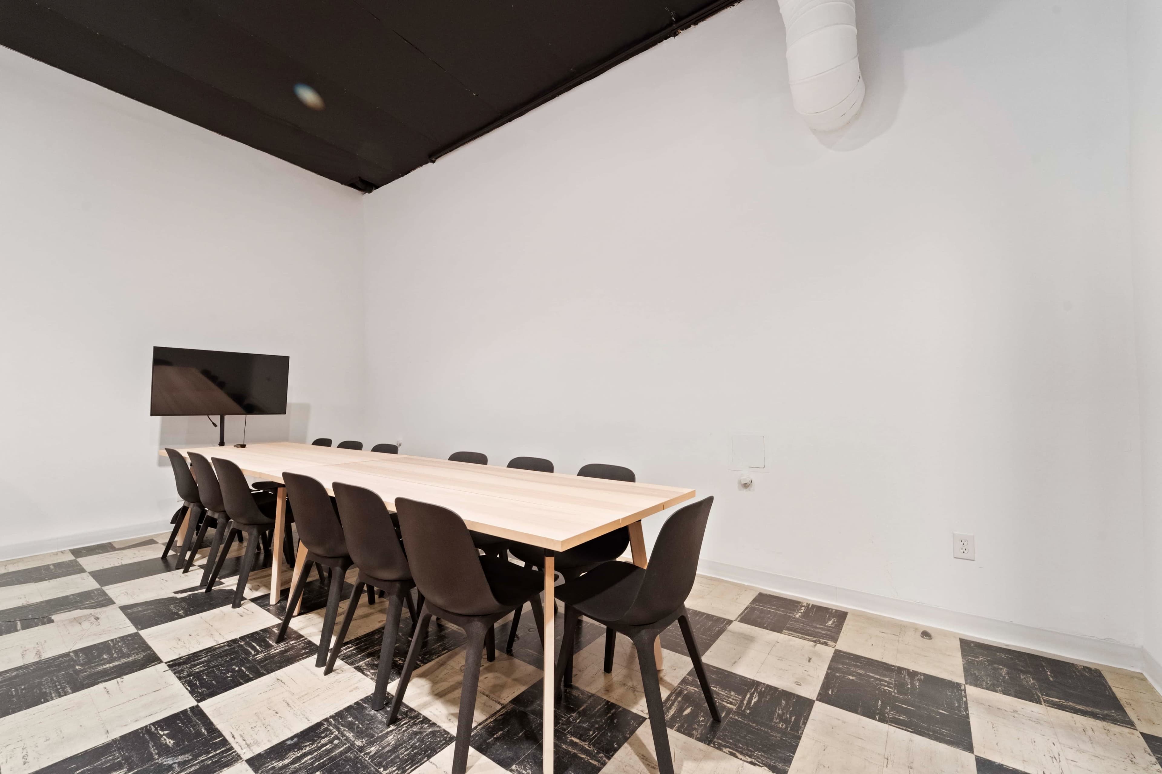 A modern meeting room with a long wooden table and black chairs, featuring a television mounted on a white wall and a checkered floor.