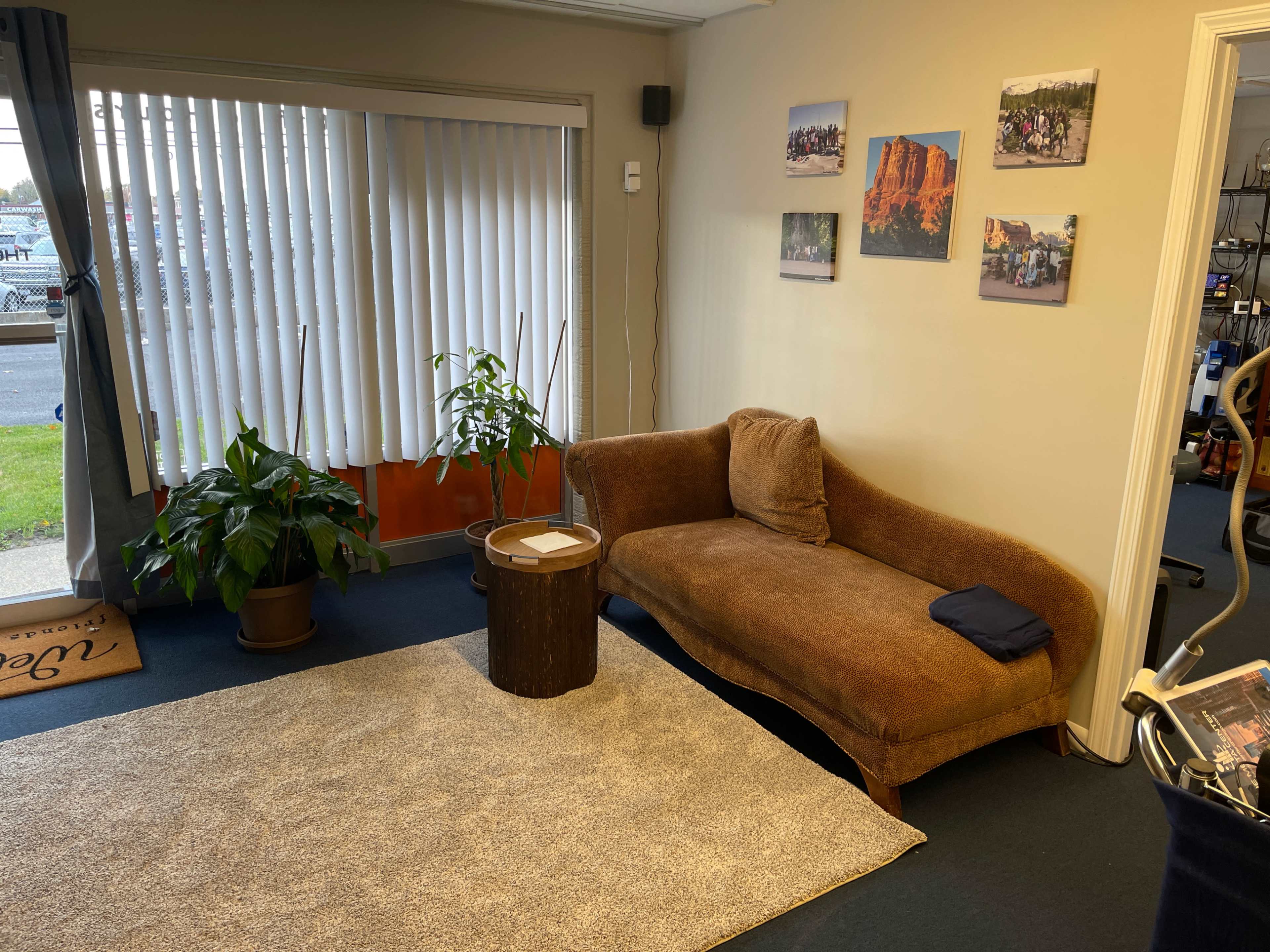 A cozy waiting area with a brown sofa, a small wooden table, a potted plant, and several framed pictures on the wall.