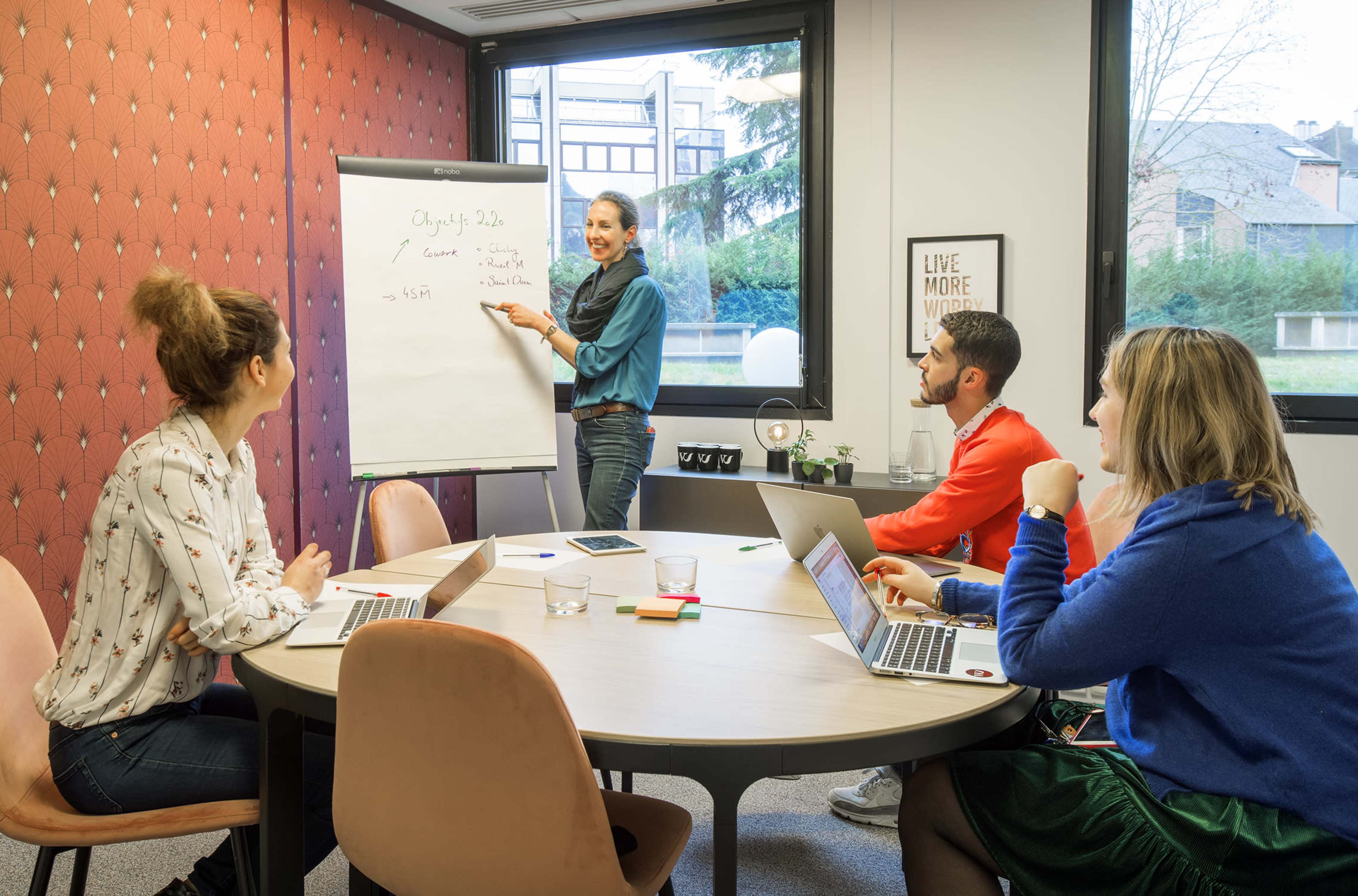 A woman presents ideas on a flip chart to three colleagues seated around a table with laptops in a bright meeting room.