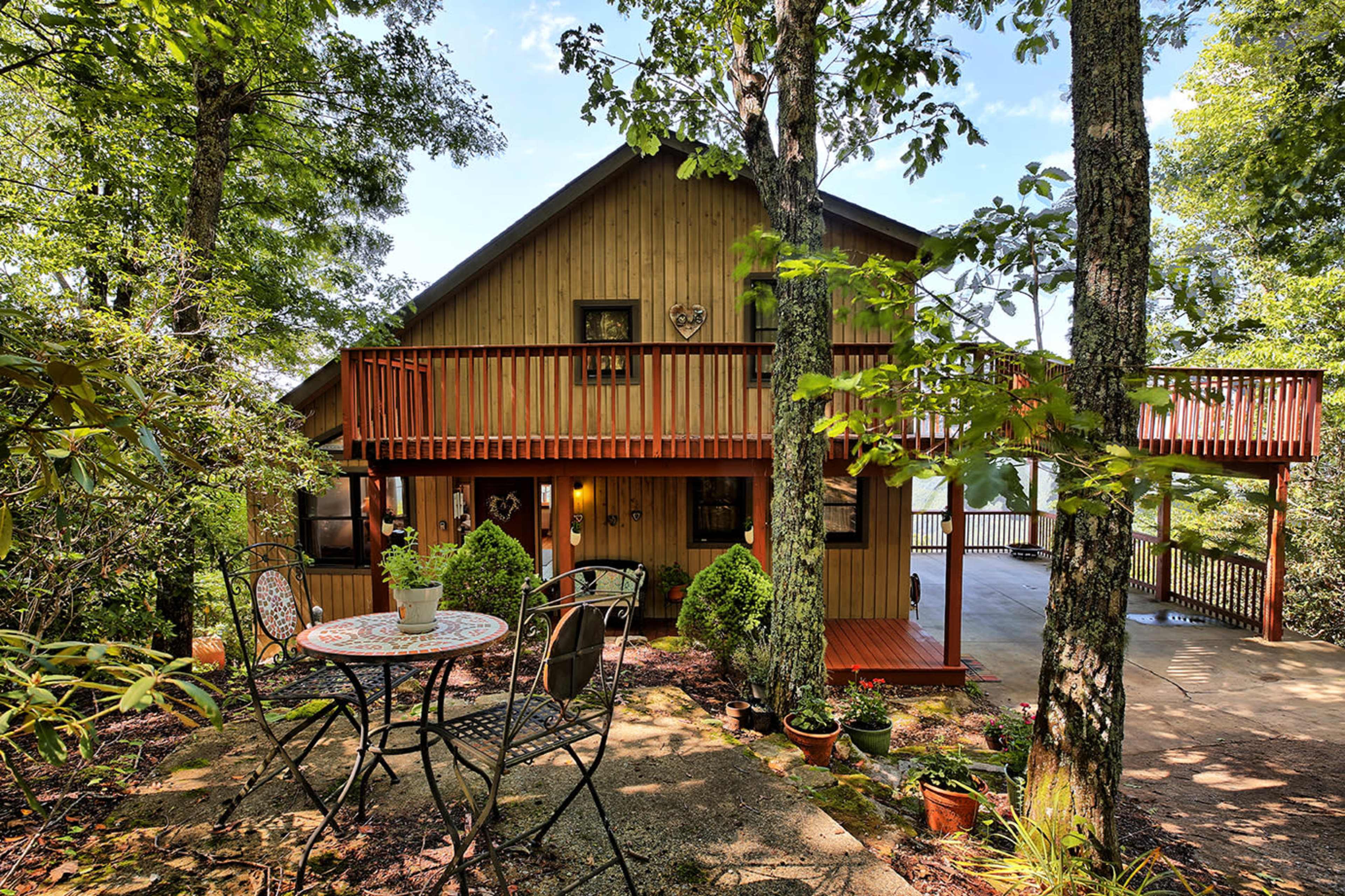 A wooden house with a balcony is surrounded by trees and features a small patio with a table and chairs in front.