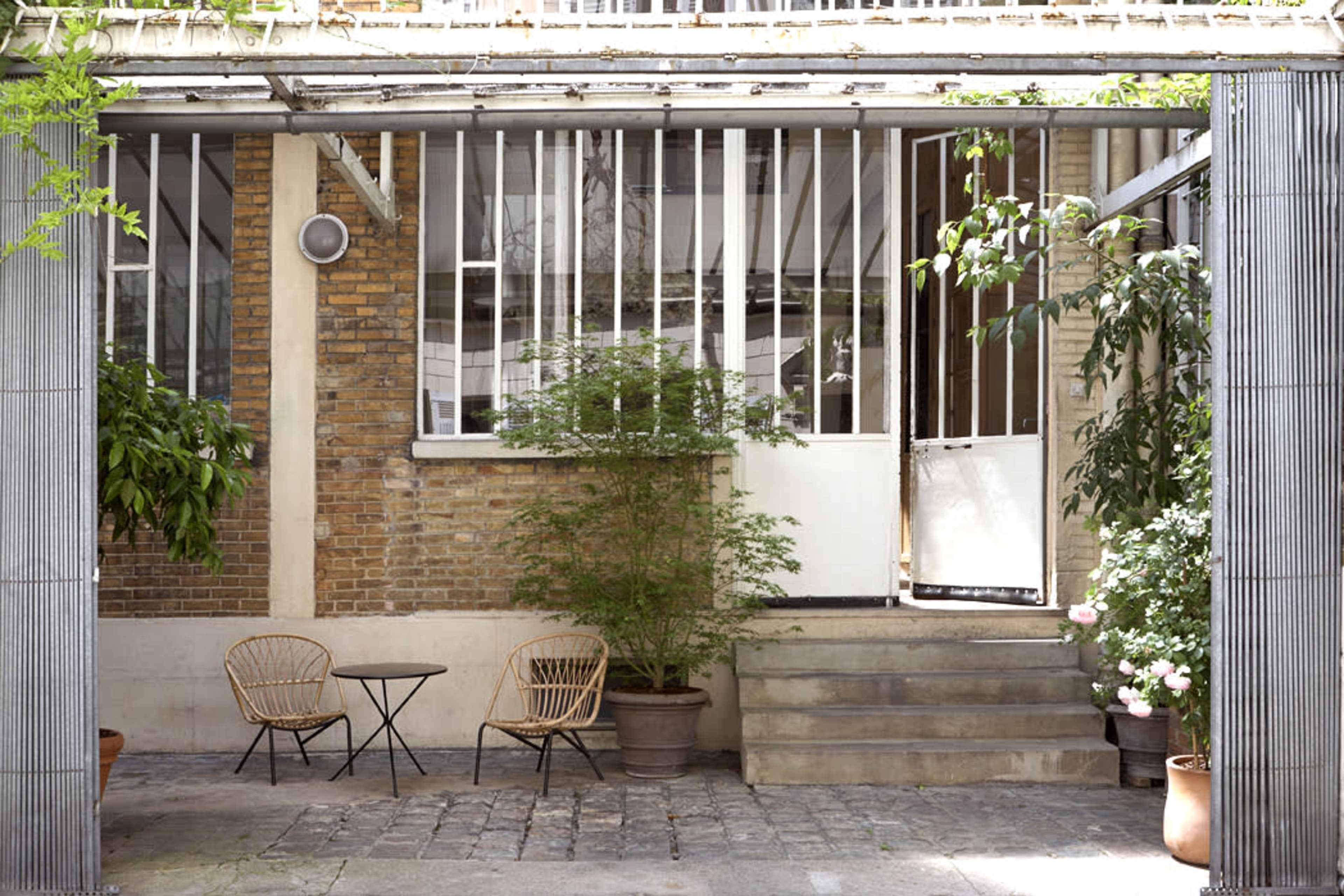 A small outdoor seating area features two chairs and a table in front of a brick wall with large windows and greenery.