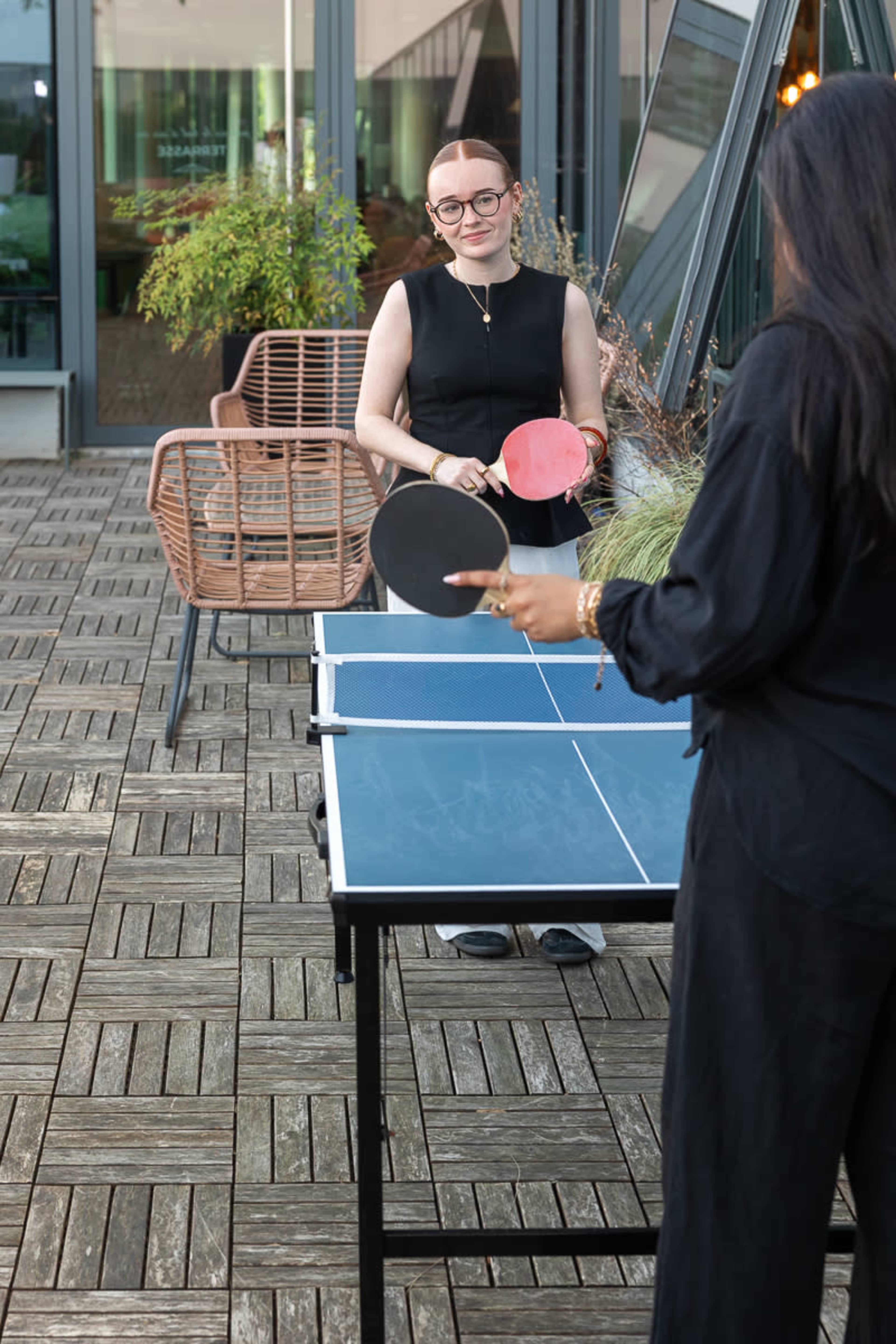 Two women are playing table tennis on an outdoor deck with modern furniture and greenery in the background.