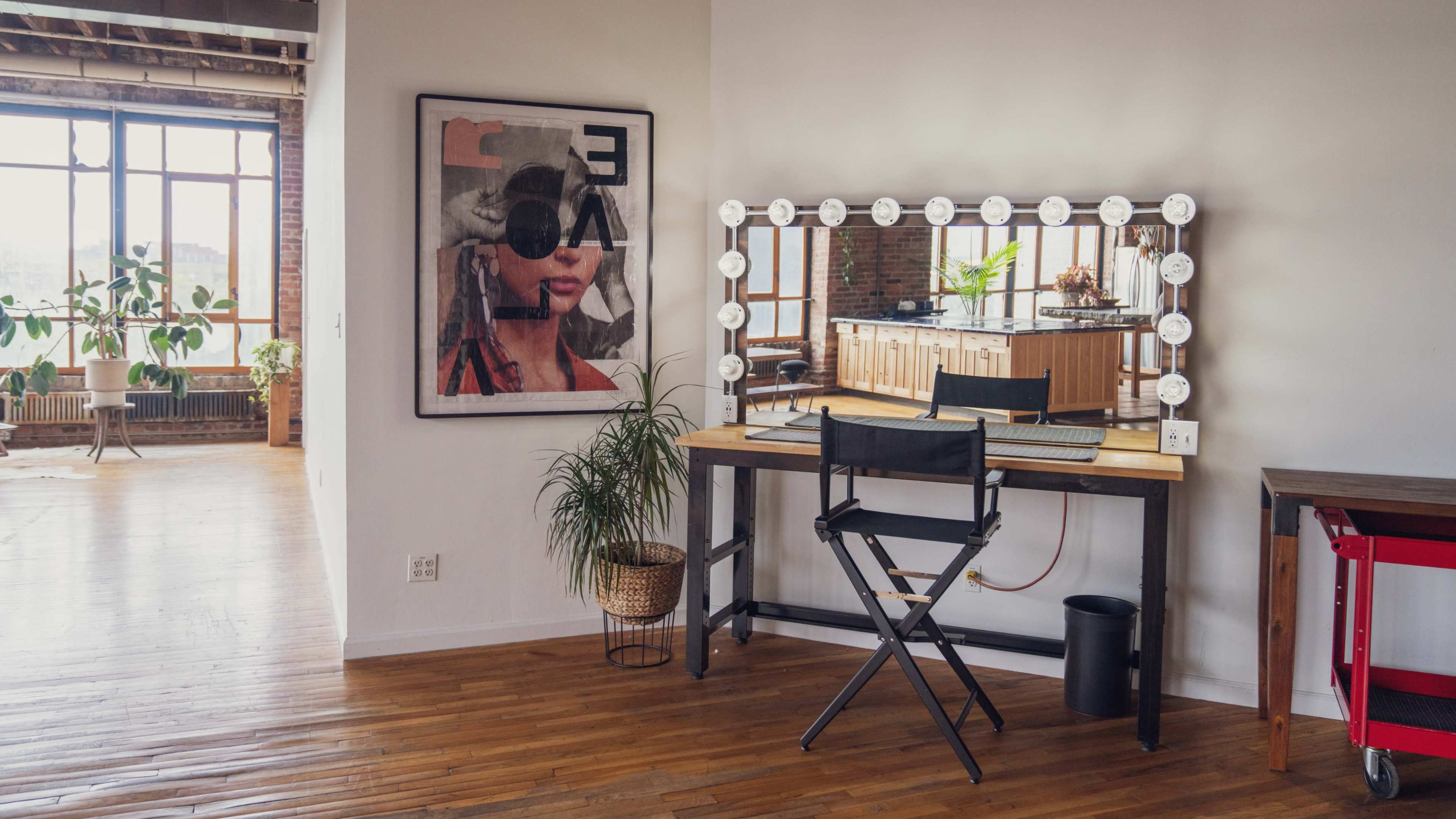 A bright room with a large window, featuring a decorated wall with a framed artwork, a vanity mirror with lights, and a director's chair next to a desk.