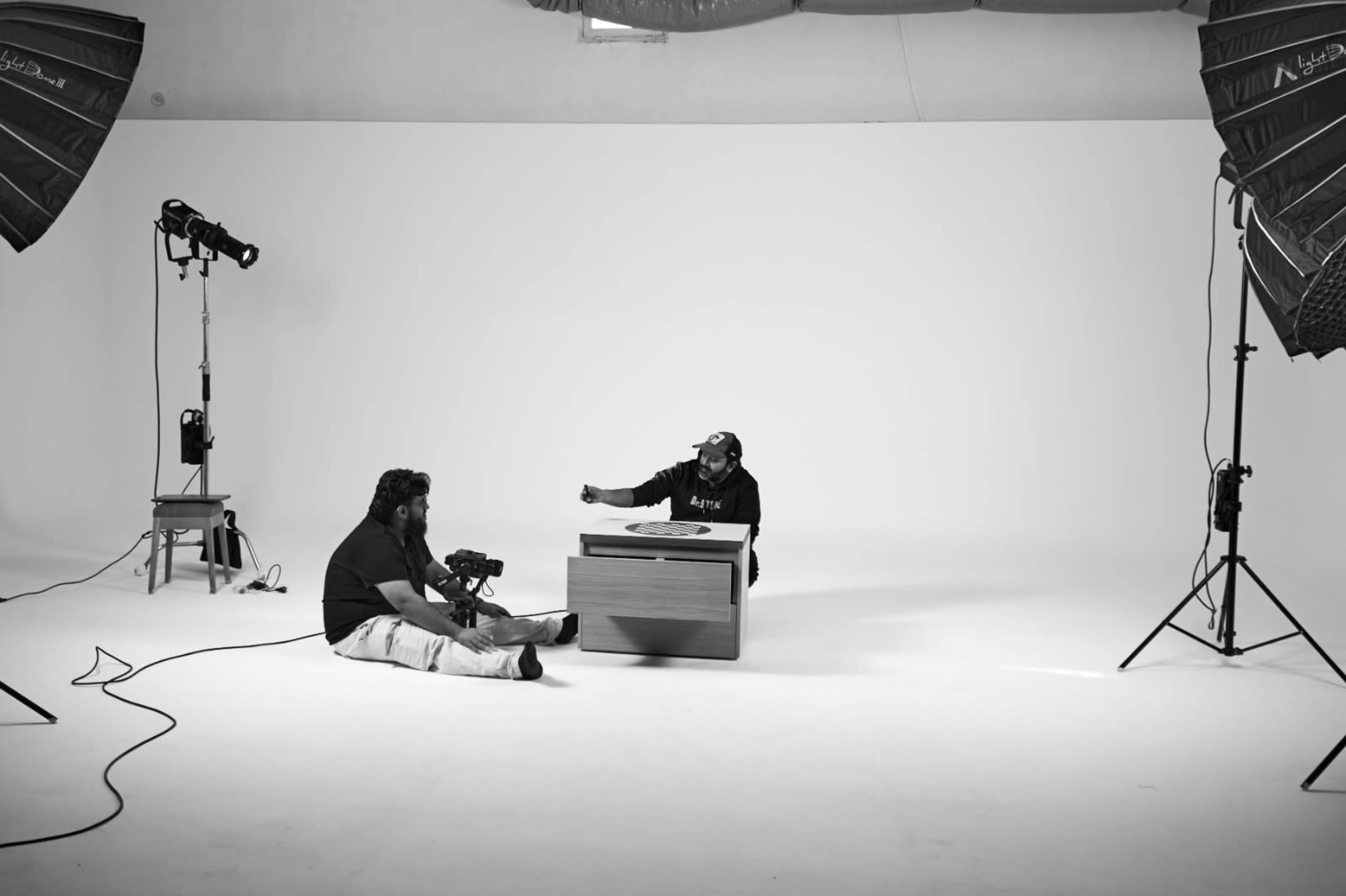 A photographer sits on the floor, aiming a camera at a person sitting at a table in a blank studio with soft lighting.