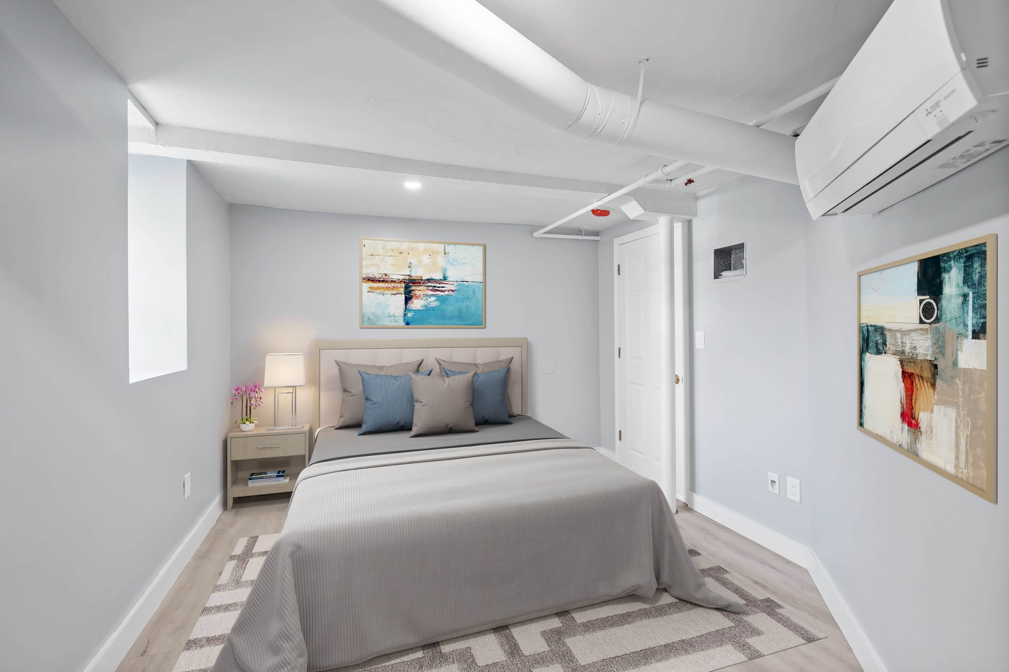 A well-lit bedroom featuring a queen-sized bed with gray bedding, light blue decorative pillows, and a small nightstand beside it.