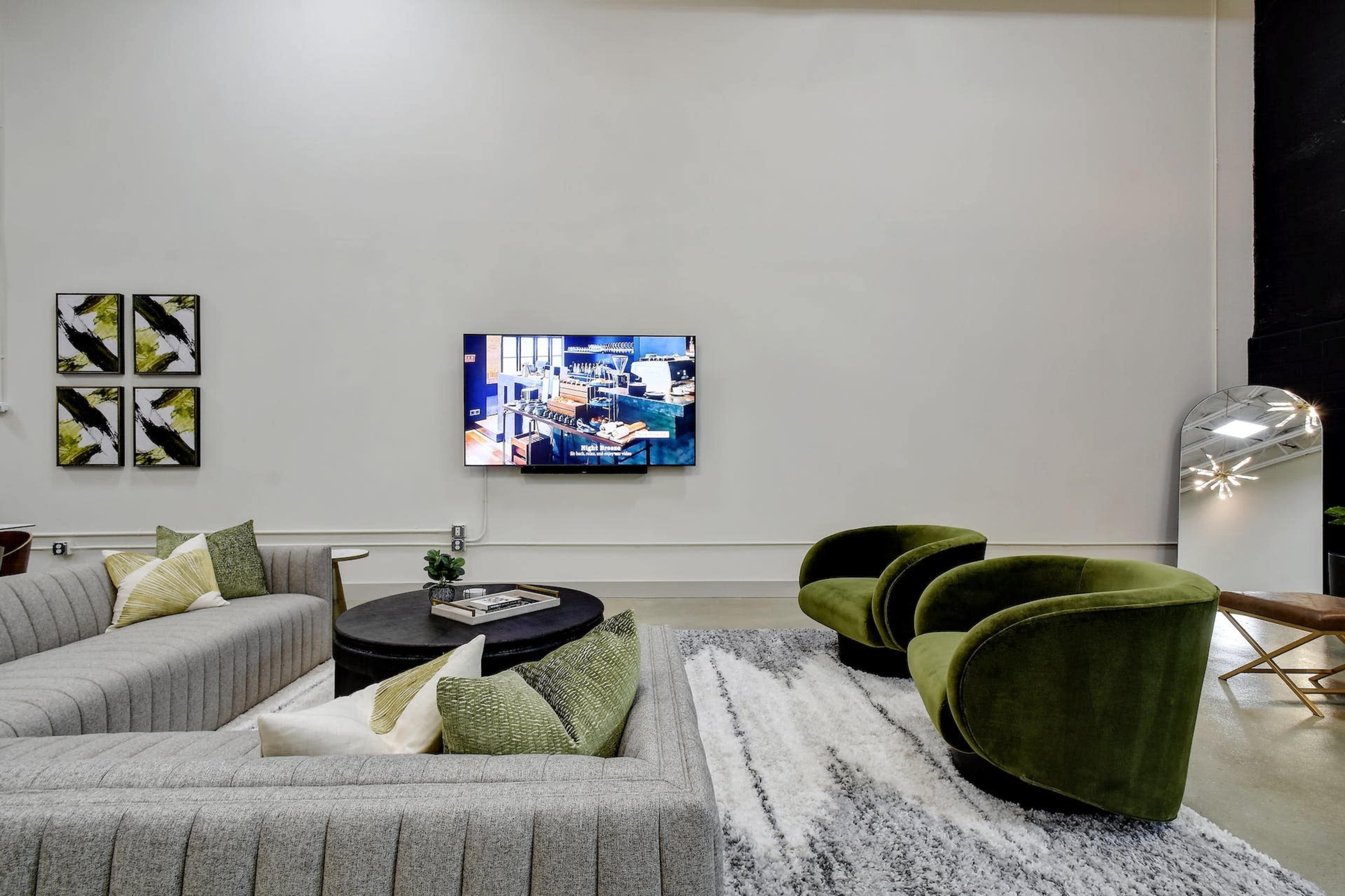 A modern living room features a gray sectional sofa, two green accent chairs, a black coffee table, a television mounted on the wall, and decorative artwork.