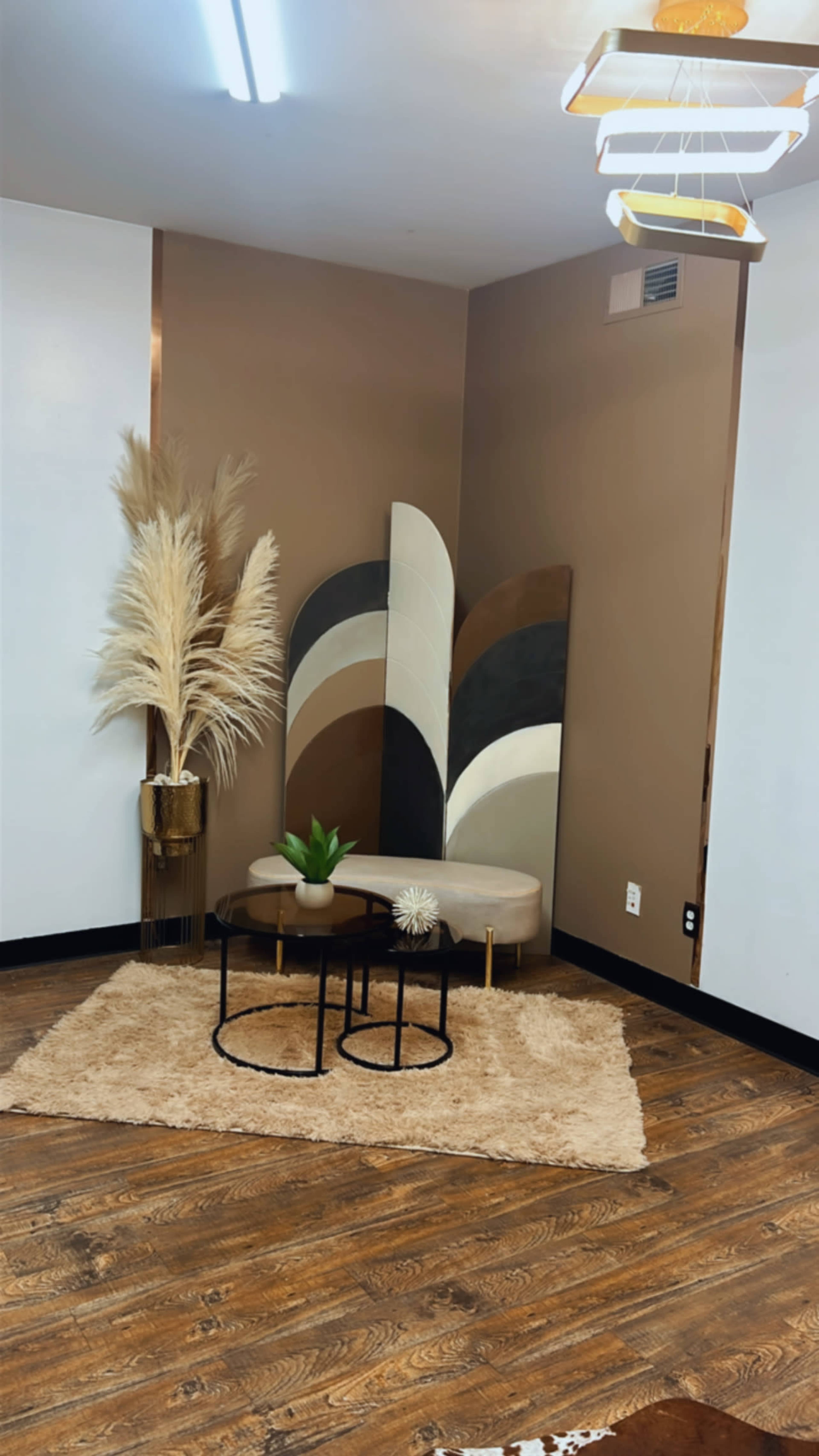 The image shows a small seating area featuring a beige couch, two black and gold side tables, a decorative plant, and a textured rug, all arranged against a backdrop of brown walls with abstract art.