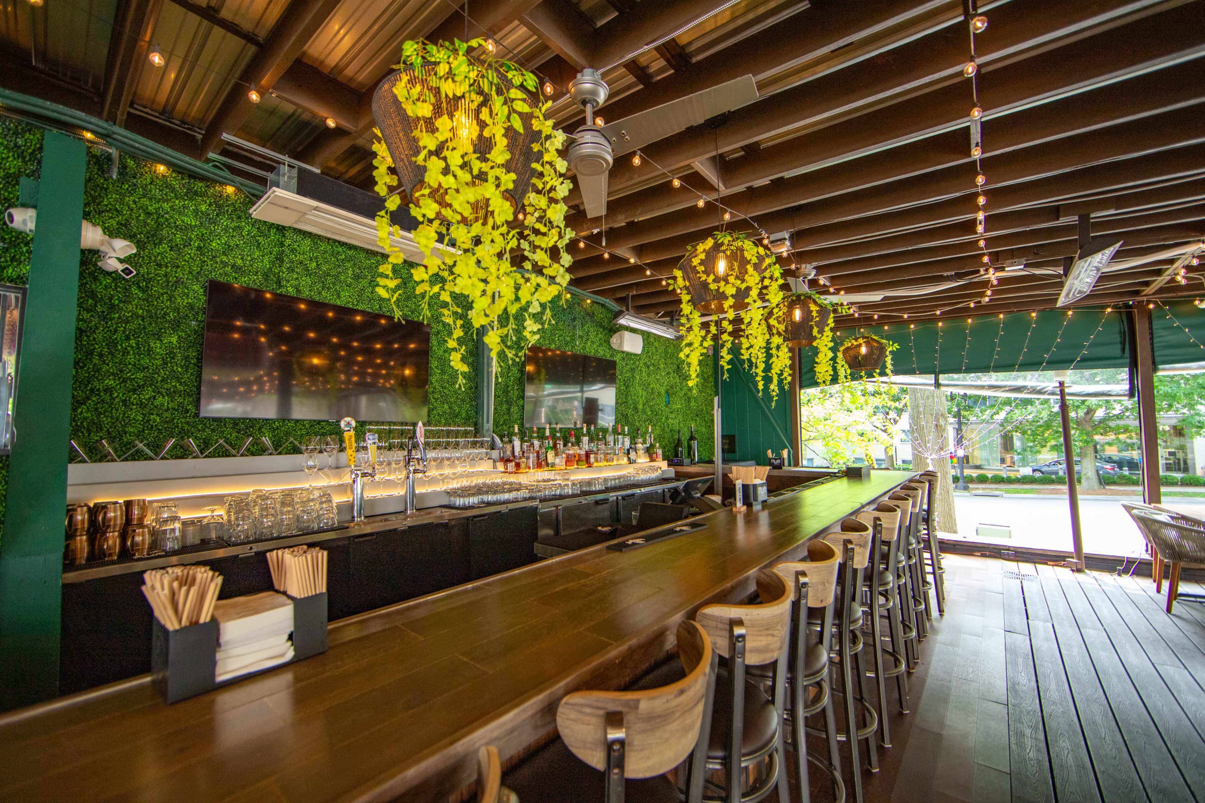A modern bar features a wooden counter, greenery on the walls, and overhead lighting with decorative plants.