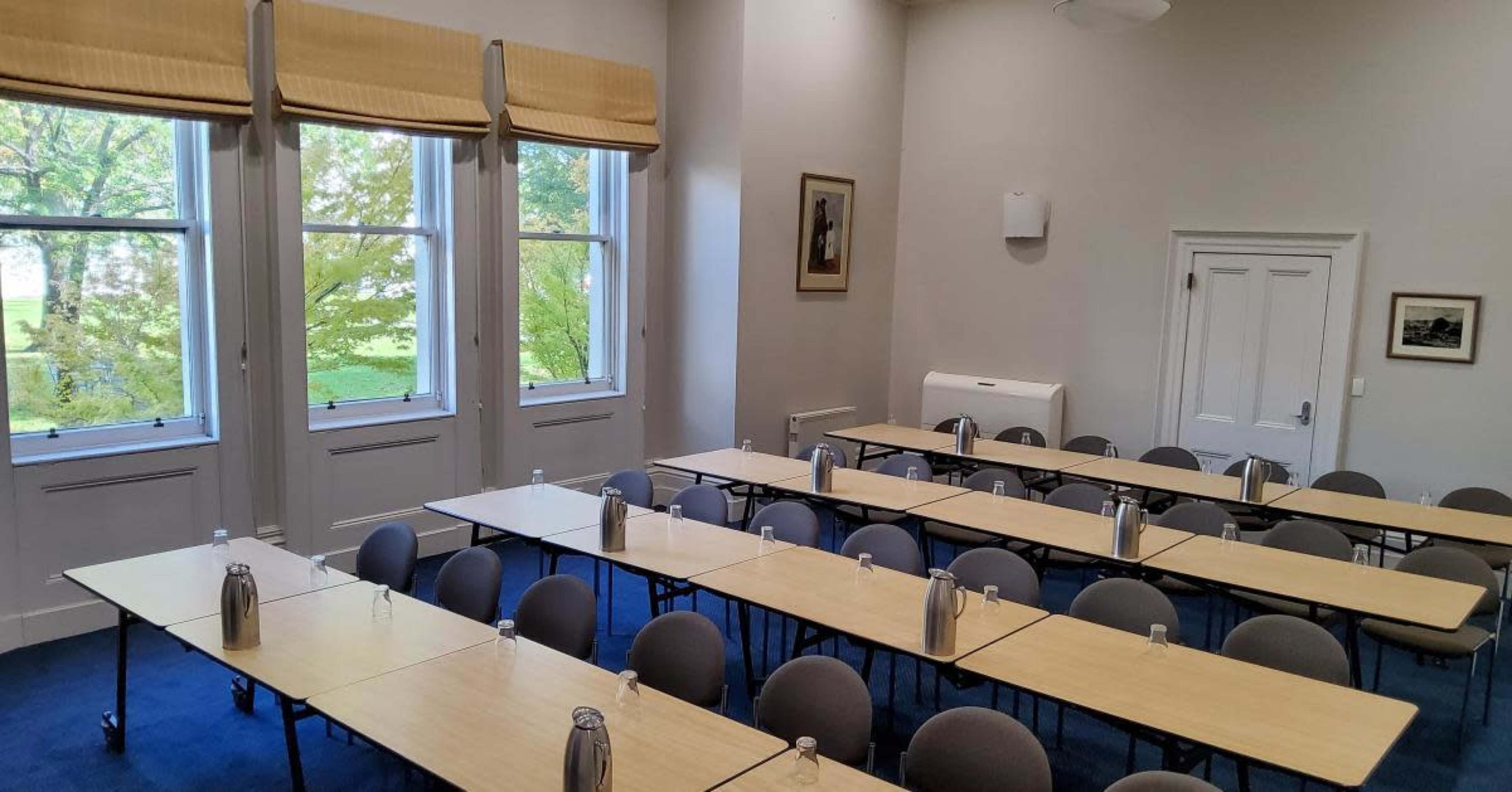 A classroom is set up with rows of tables and chairs, each equipped with a water bottle, and large windows provide a view of greenery outside.