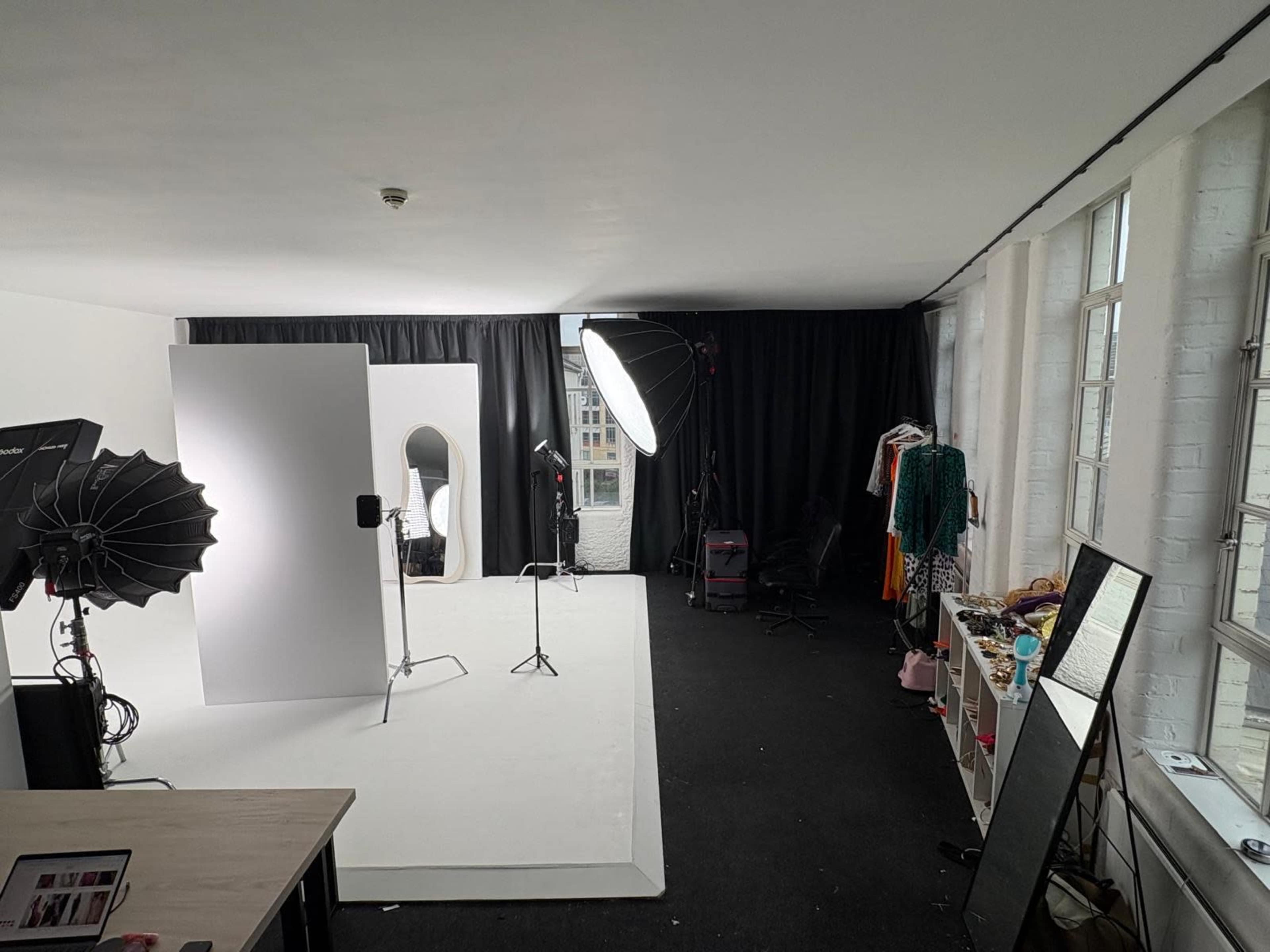 The image shows a photography studio with softbox lights, a blank backdrop, a mirror, and various clothing items arranged along the walls.