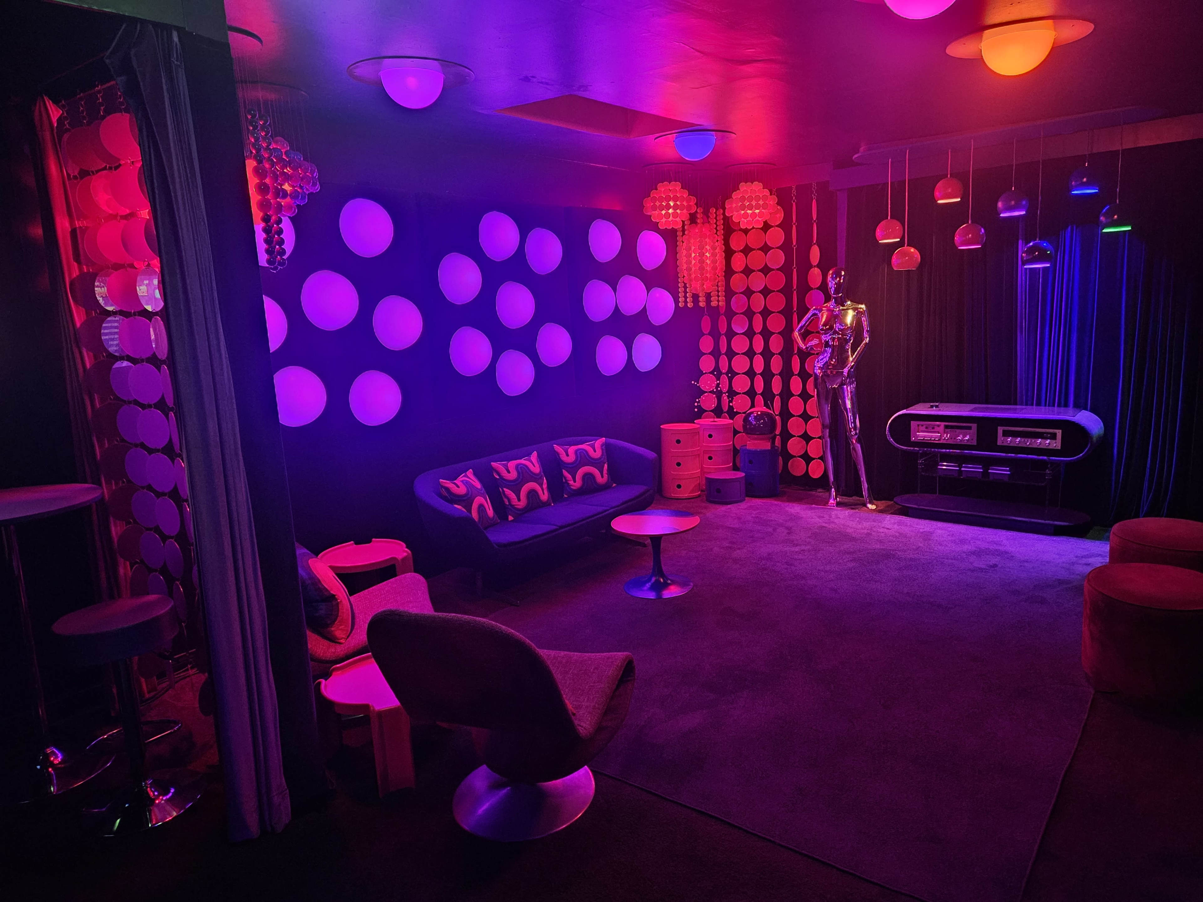The scene features a vibrant retro-themed lounge with colorful lighting, a mix of modern furniture, and circular wall decorations.