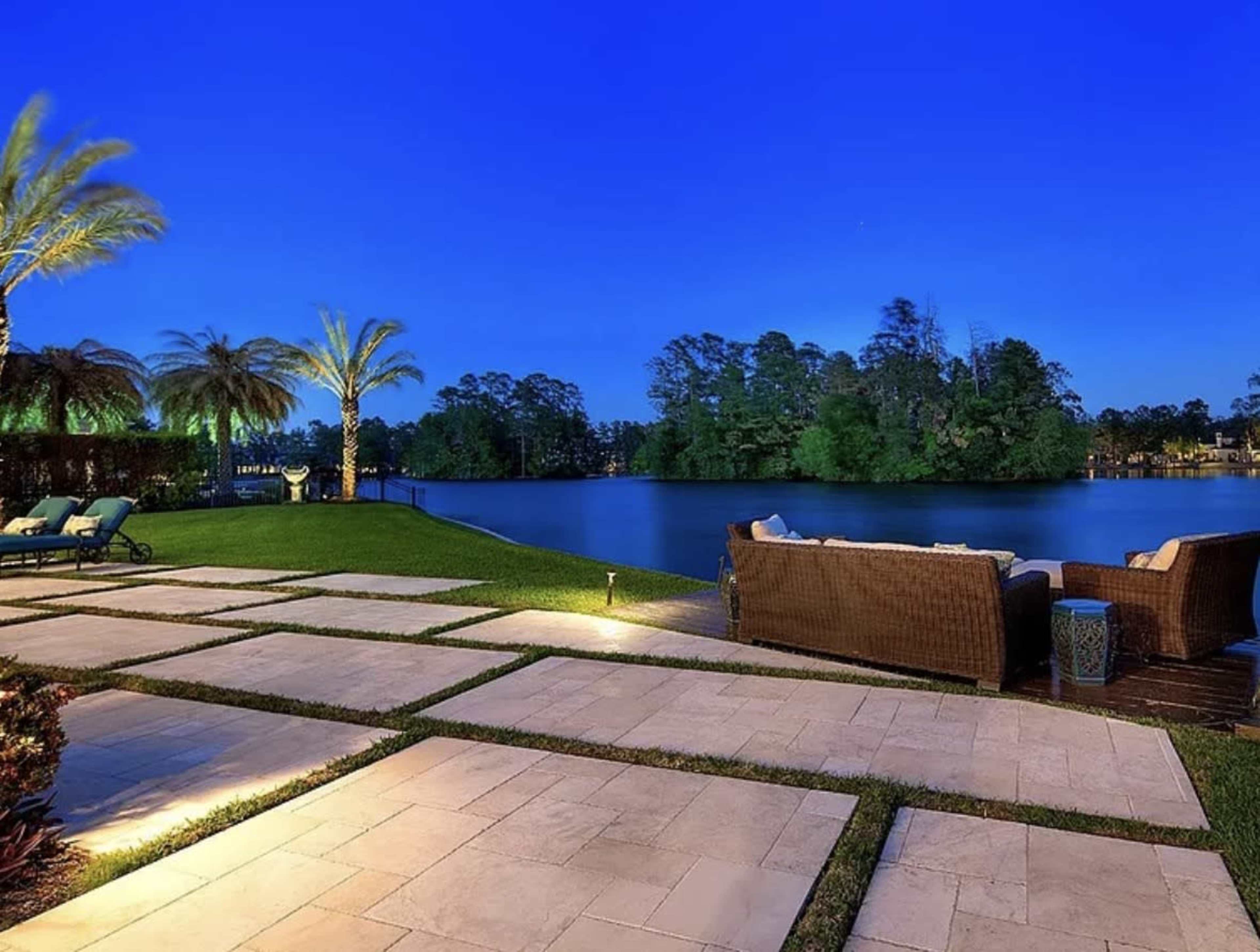 The image shows a peaceful outdoor patio area with seating, palm trees, and a calm lake under a twilight sky.