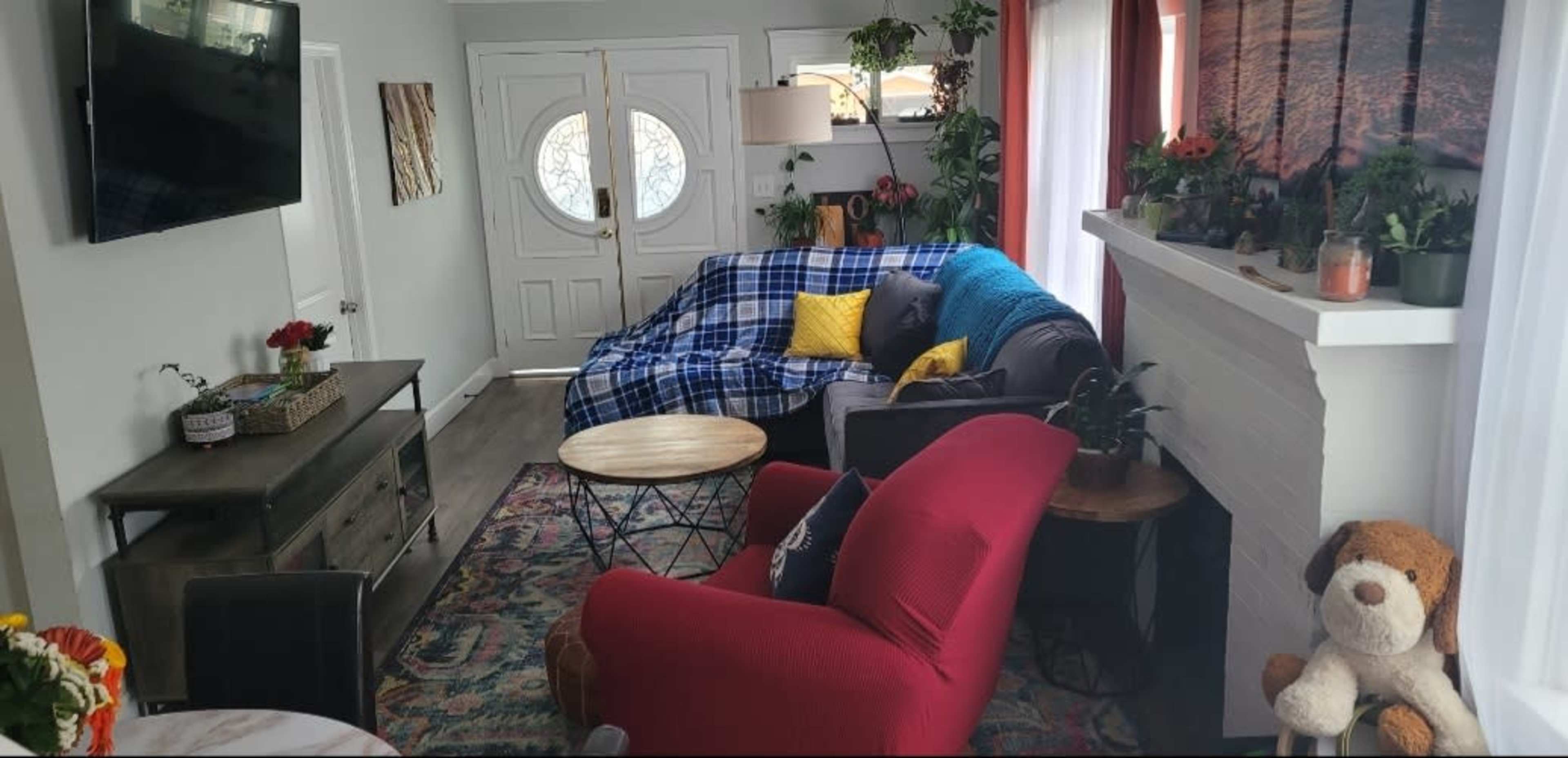 The image shows a living room with a couch covered in a blue and plaid blanket, a red armchair, a coffee table, and decorative plants, all arranged around a central area.