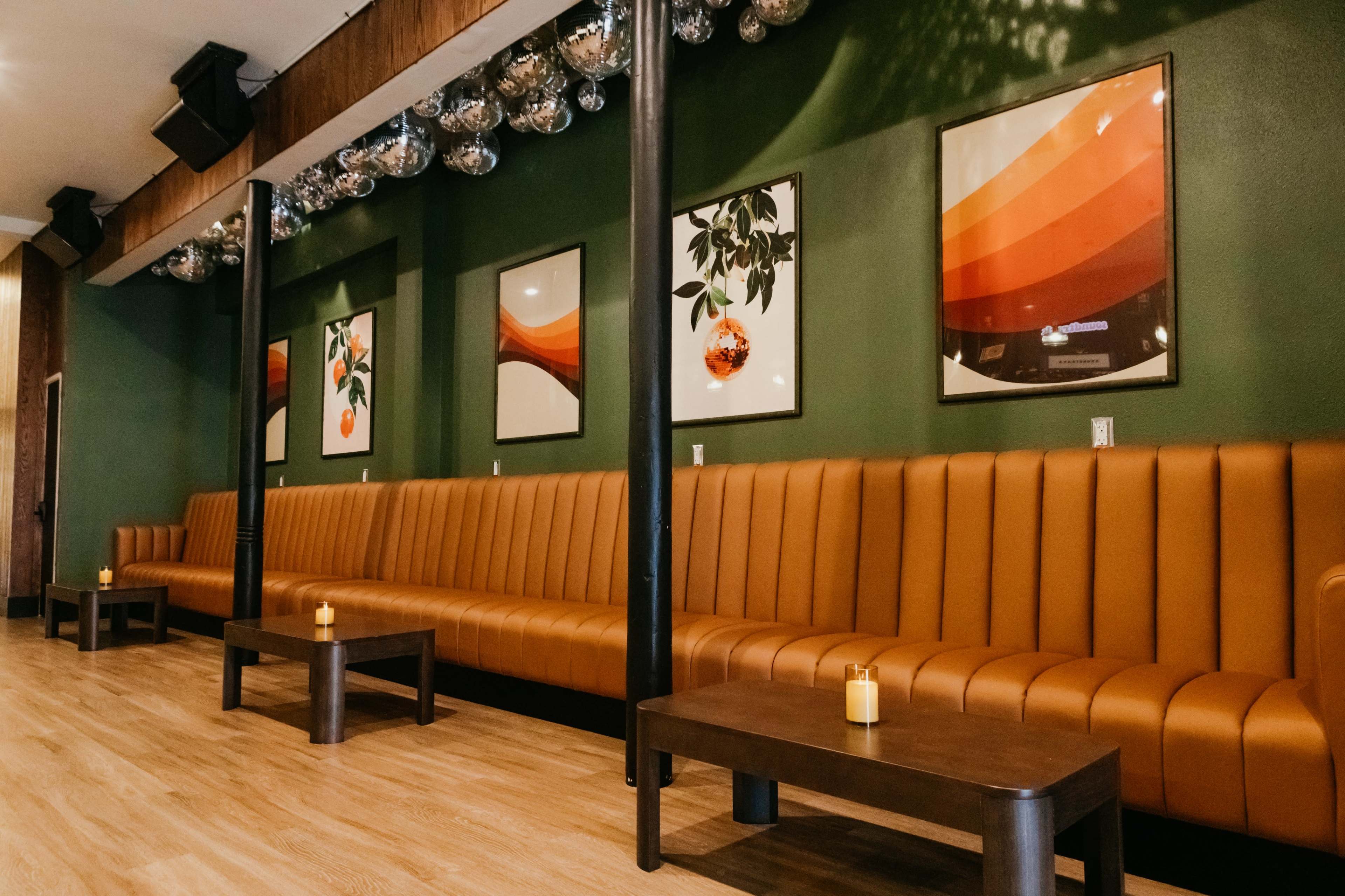 The image shows a lounge area with a long brown leather bench, dark wooden tables, and framed art on green walls, illuminated by soft lighting.