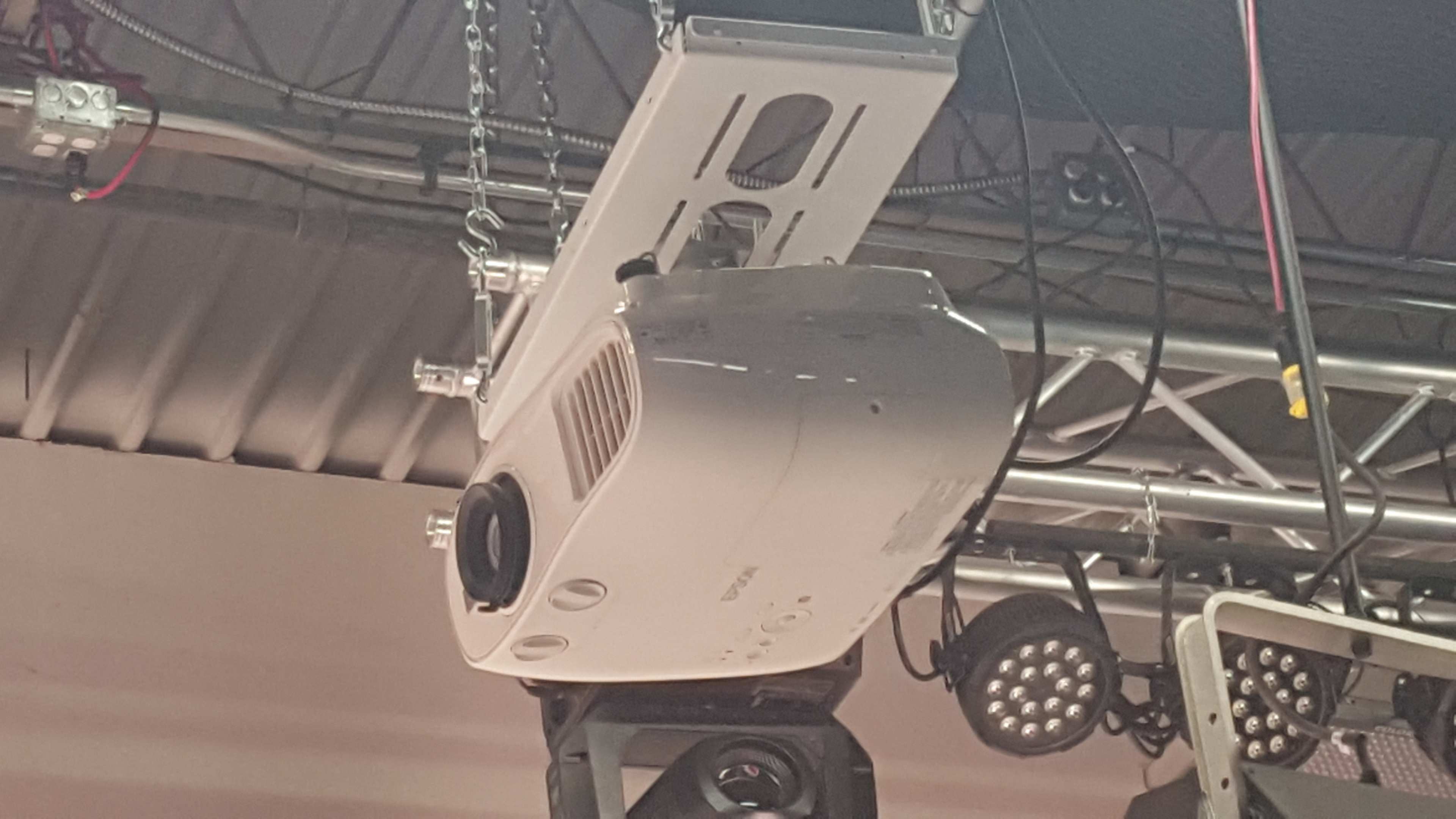 A ceiling-mounted projector is suspended from a truss above a stage area, with various light fixtures in the background.