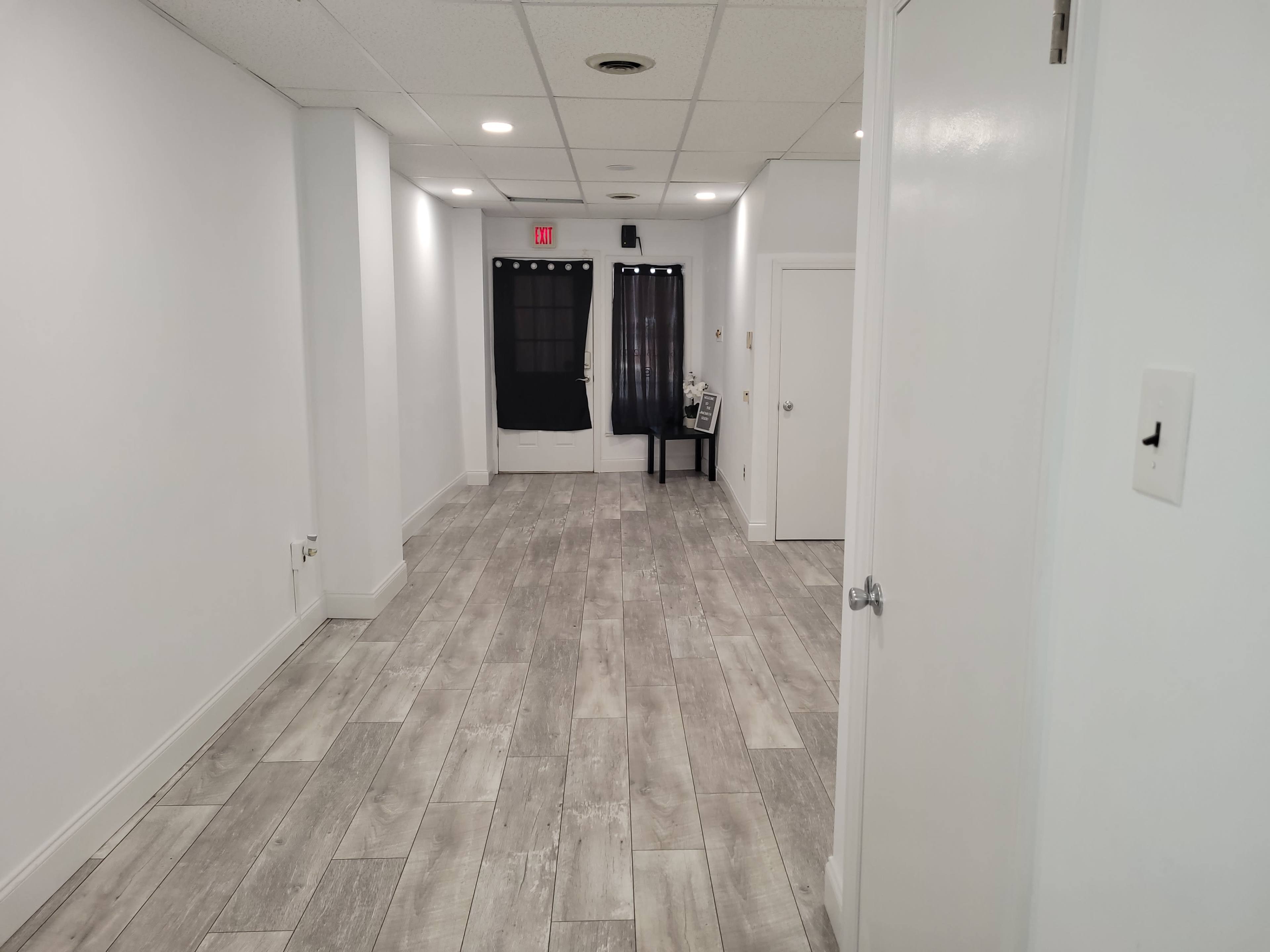 The image shows a long, narrow hallway with grey tiled flooring and two black curtains at the end, alongside a door and a small table.