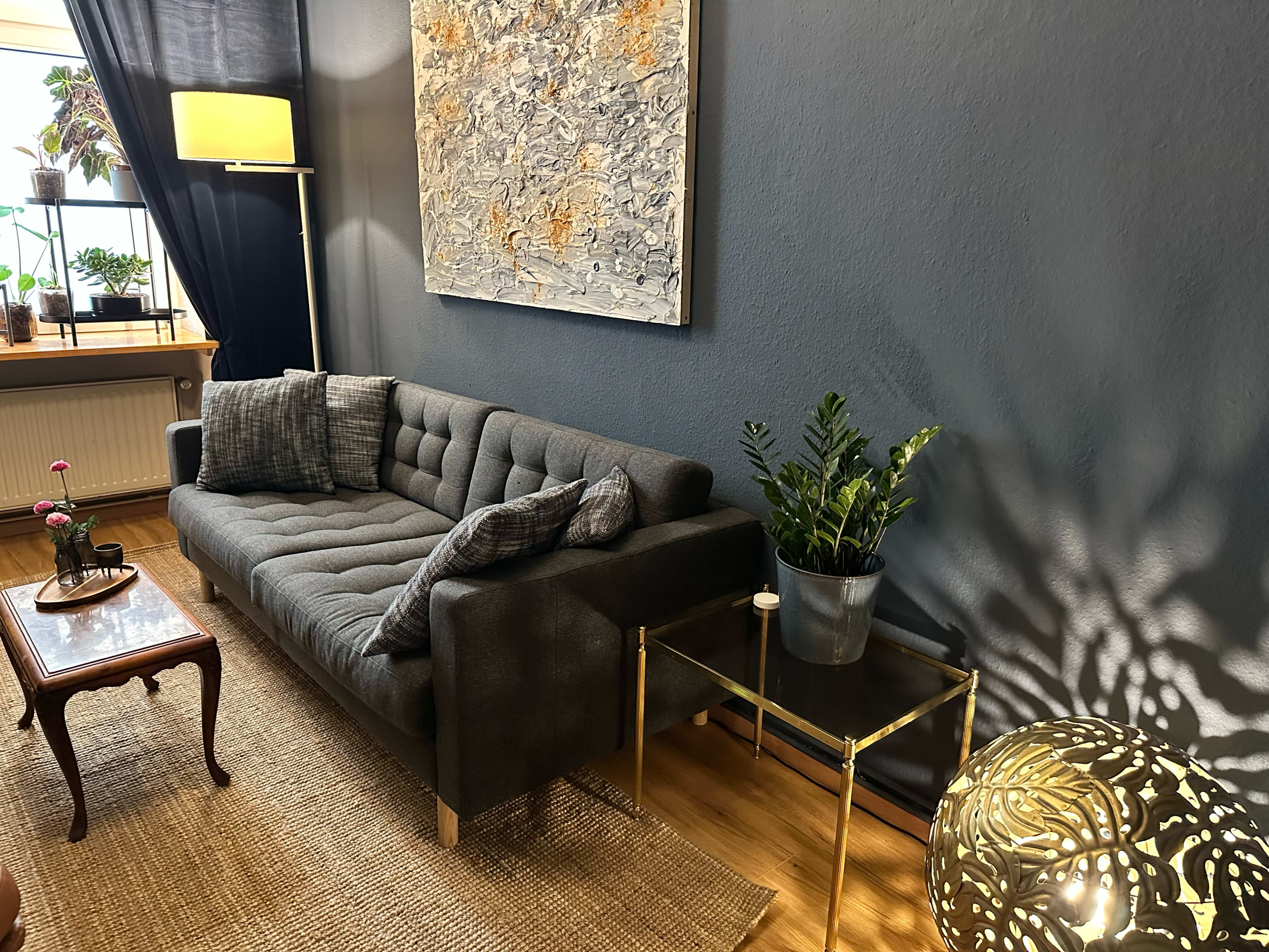 The image shows a modern living room with a dark gray sofa, a small side table with a plant, and decorative artwork on the wall.