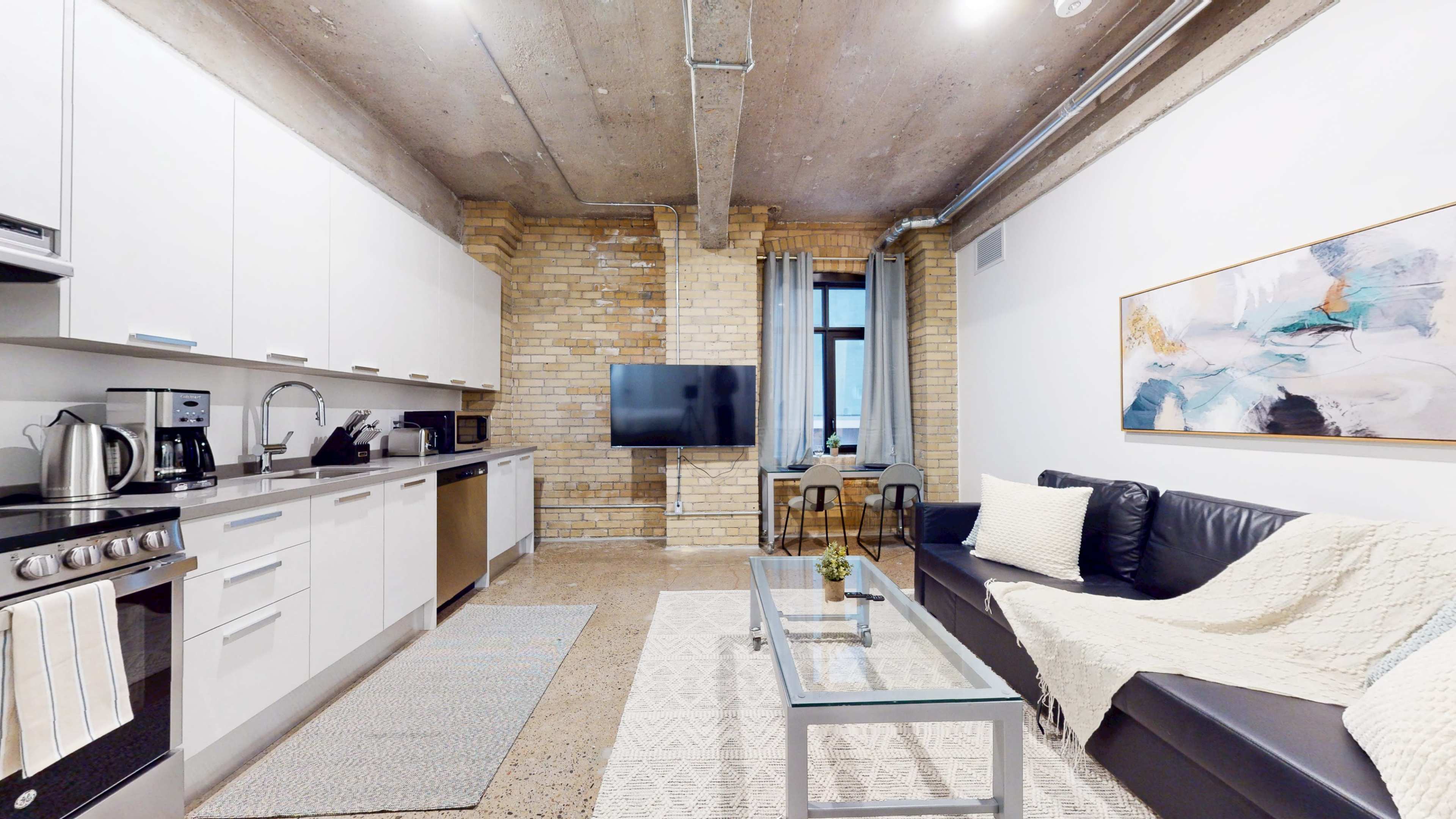 The image shows a modern apartment interior featuring a kitchen area on the left, a living space with a black sofa and coffee table in the center, and a television mounted on a brick wall.