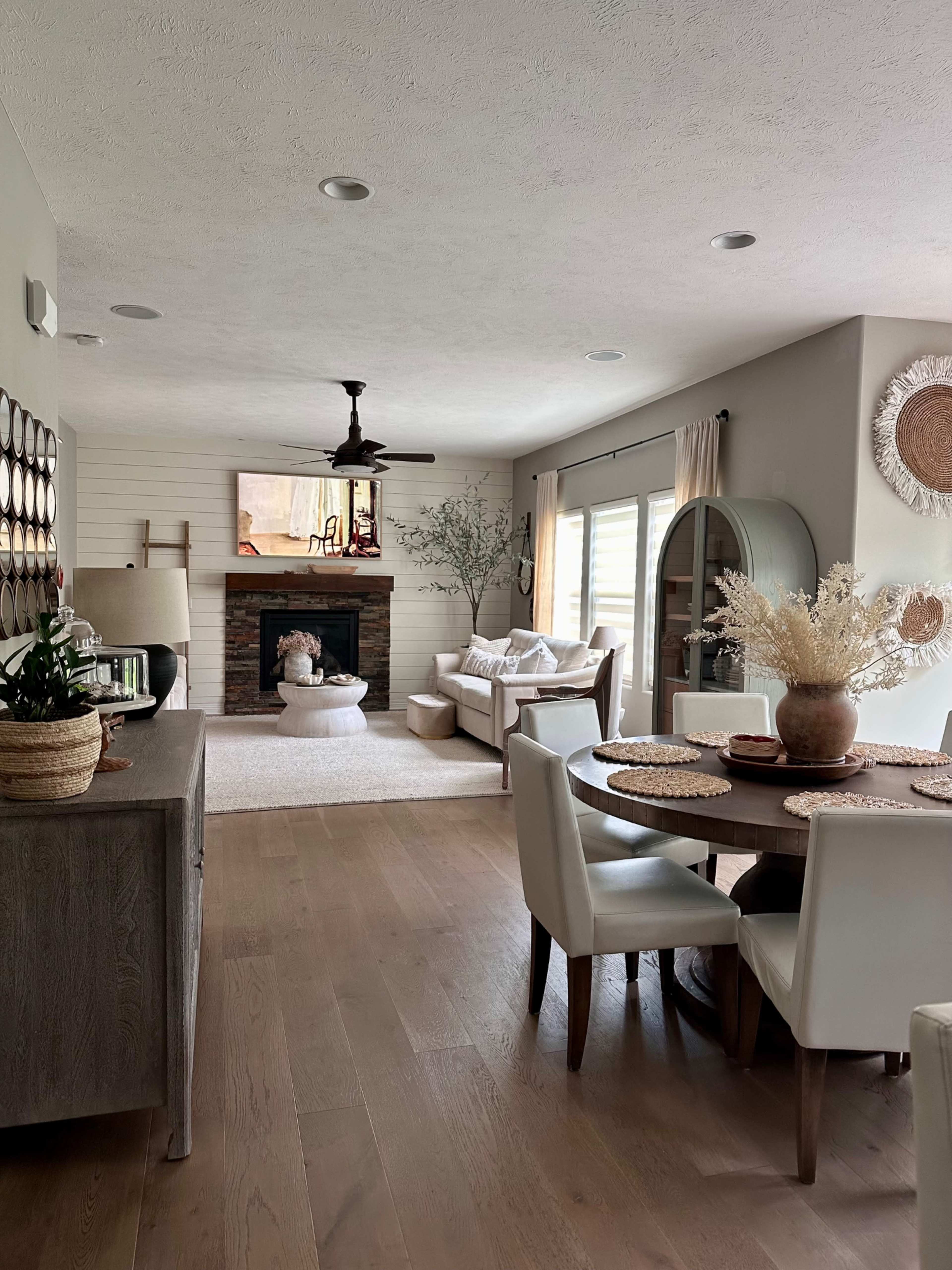The image shows a modern living space featuring a dining area with a round table and chairs, a cozy living room with a fireplace, and warm, neutral decor throughout.