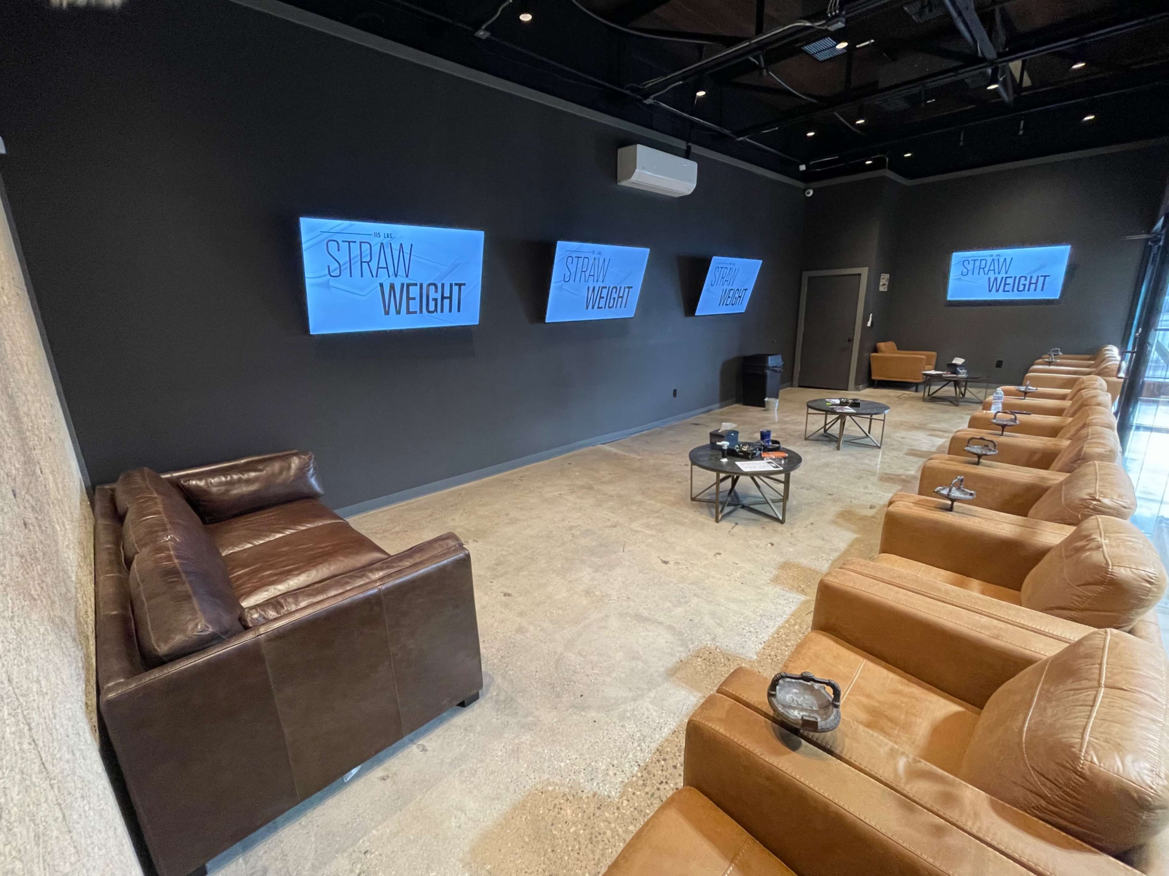A modern waiting area features brown leather seating, large screens displaying "STRAW WEIGHT," and minimalist decor.