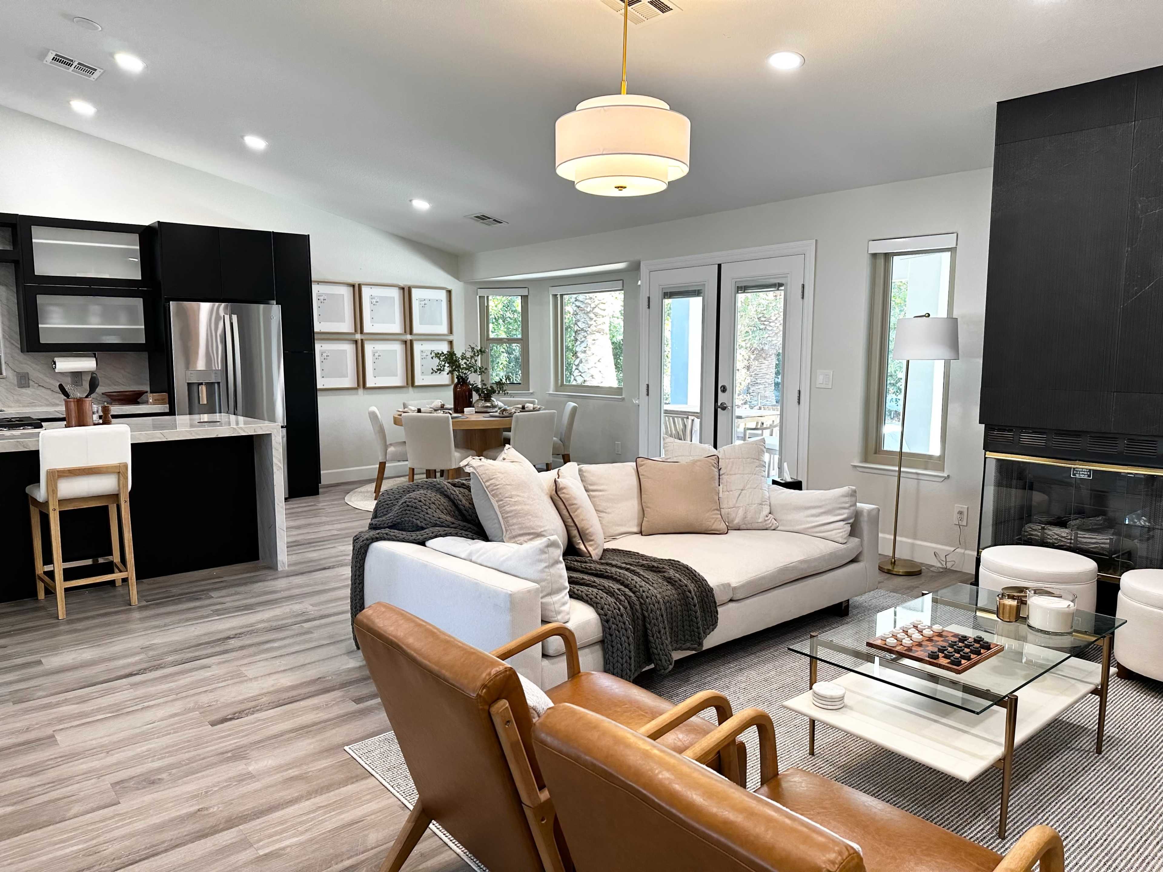 A modern living room features a white sectional sofa, brown leather chairs, a glass coffee table, and an open kitchen area in the background with seating and a dining table.