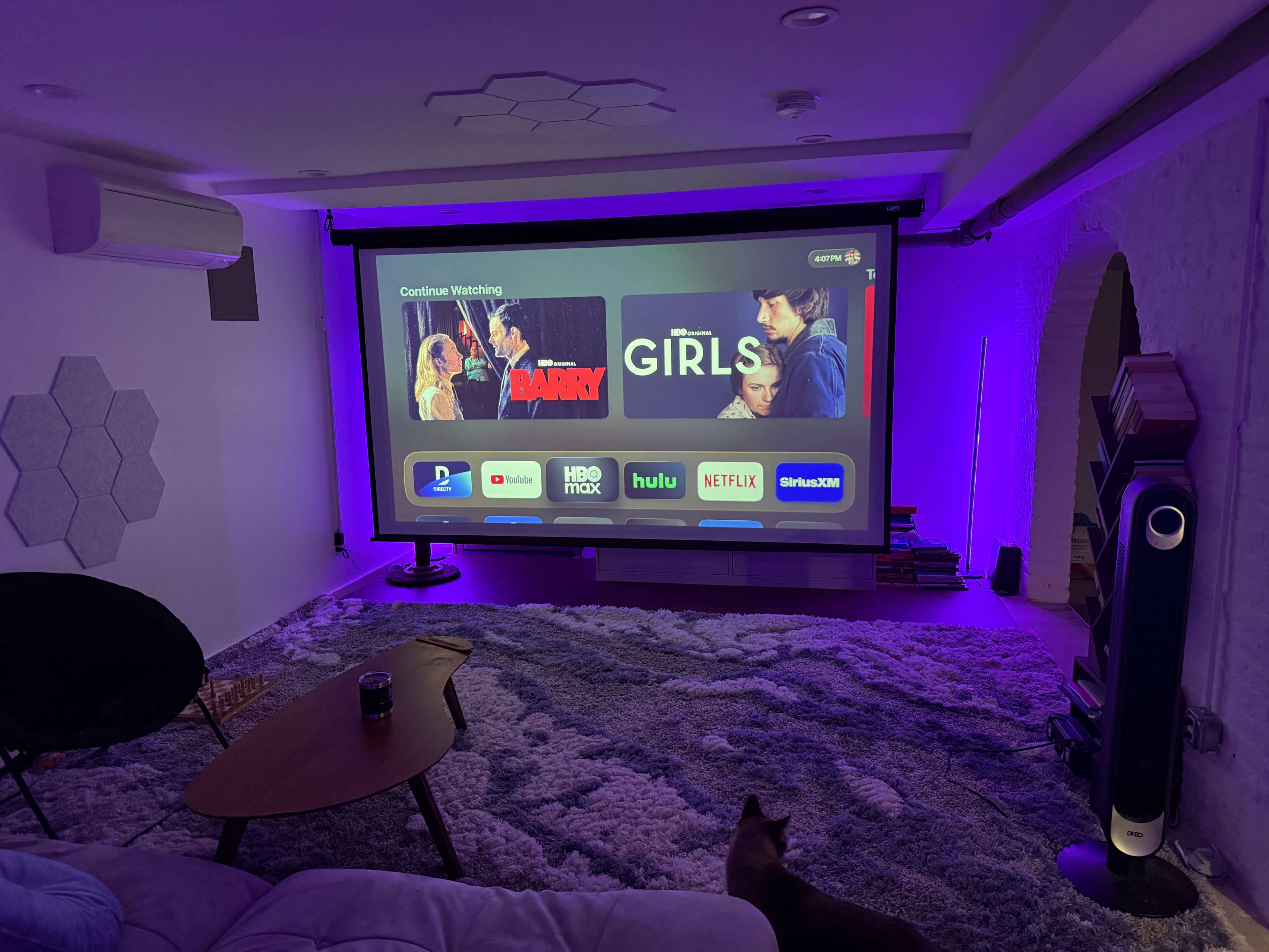 A large projector screen displays streaming options for various shows in a cozy living room with purple ambient lighting and a plush rug.