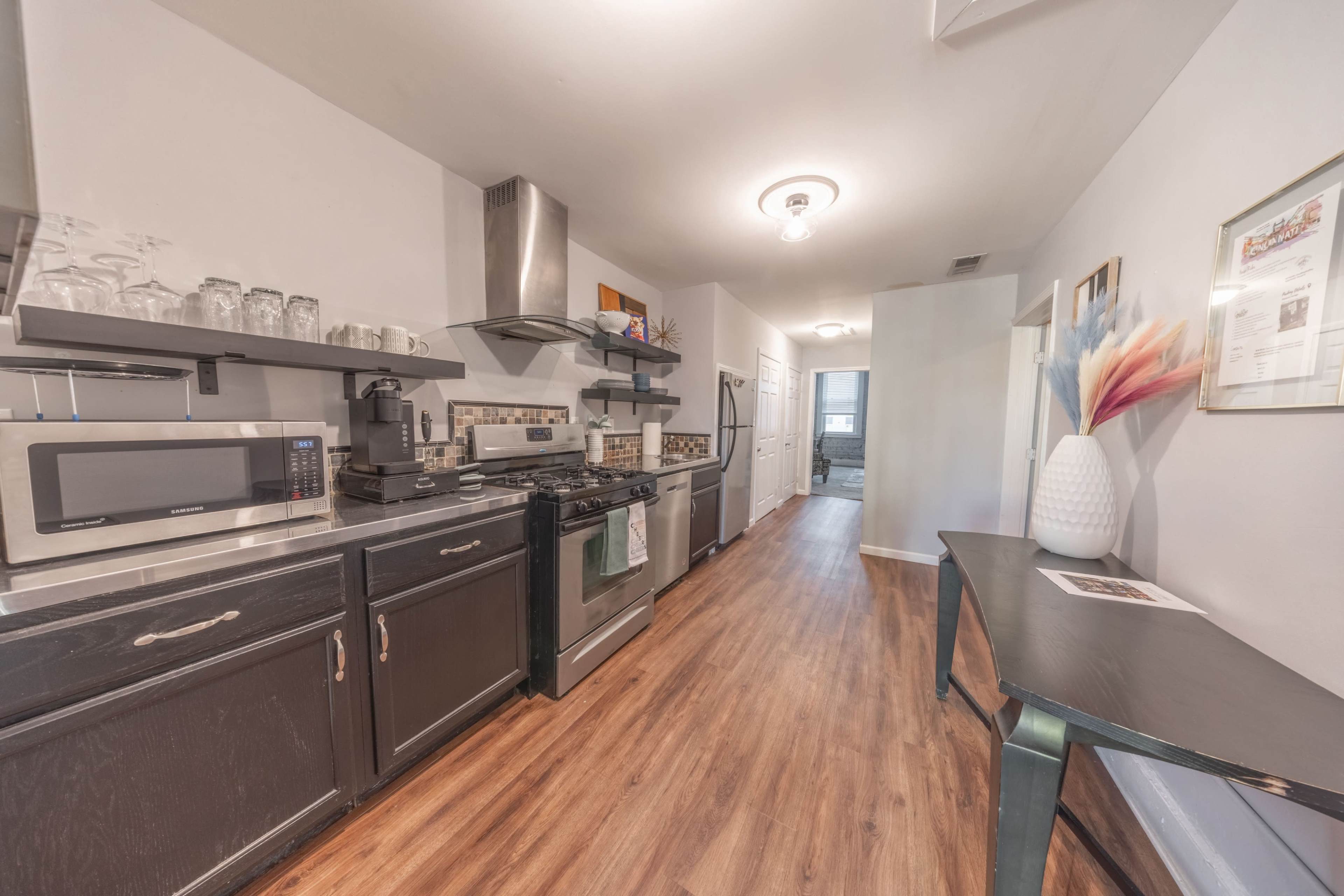 The image shows a modern kitchen with stainless steel appliances, dark cabinets, and a hardwood floor, leading to a hallway.