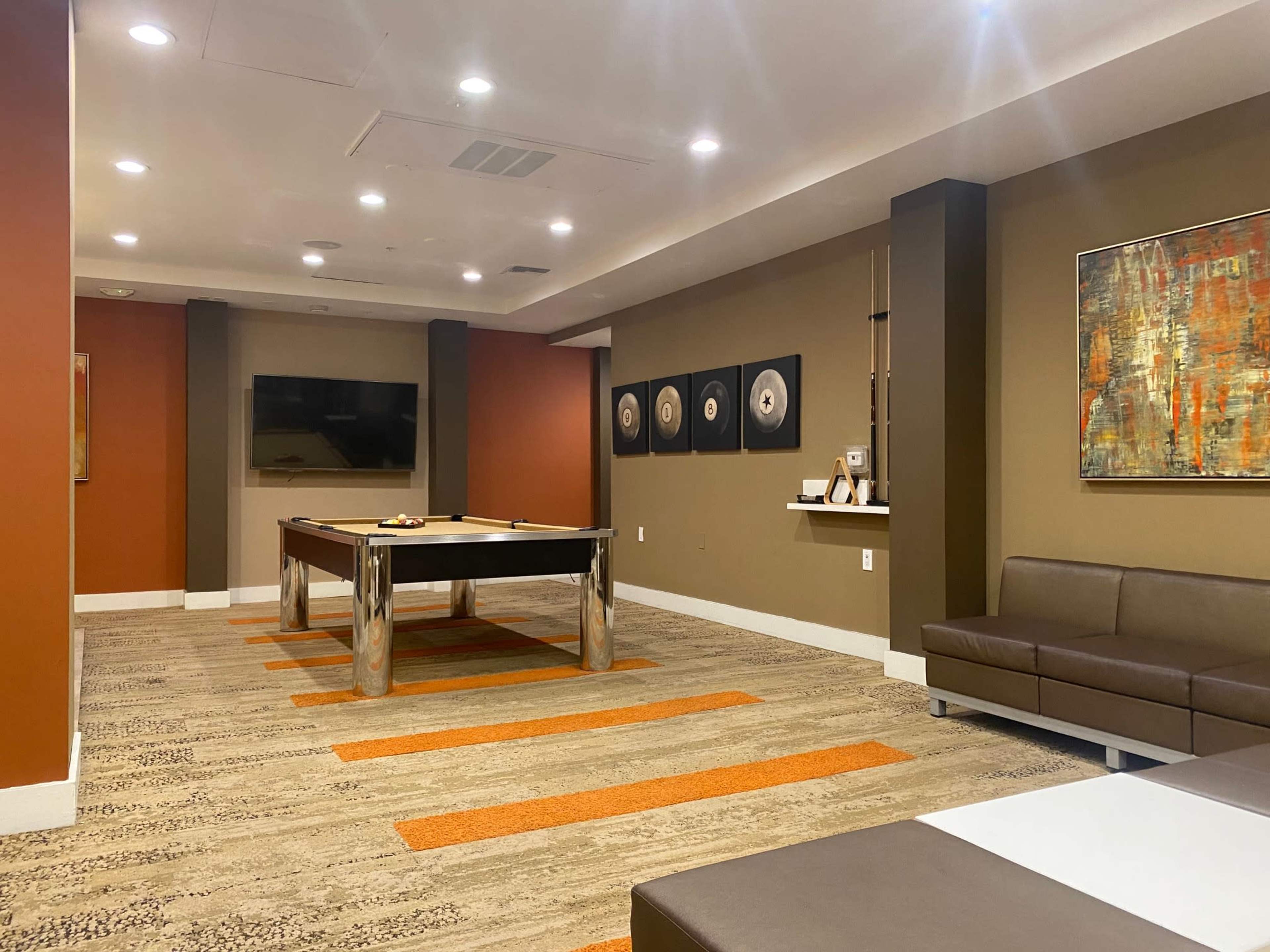 A room featuring a pool table, a television on the wall, and several framed artworks, with orange and beige decor.