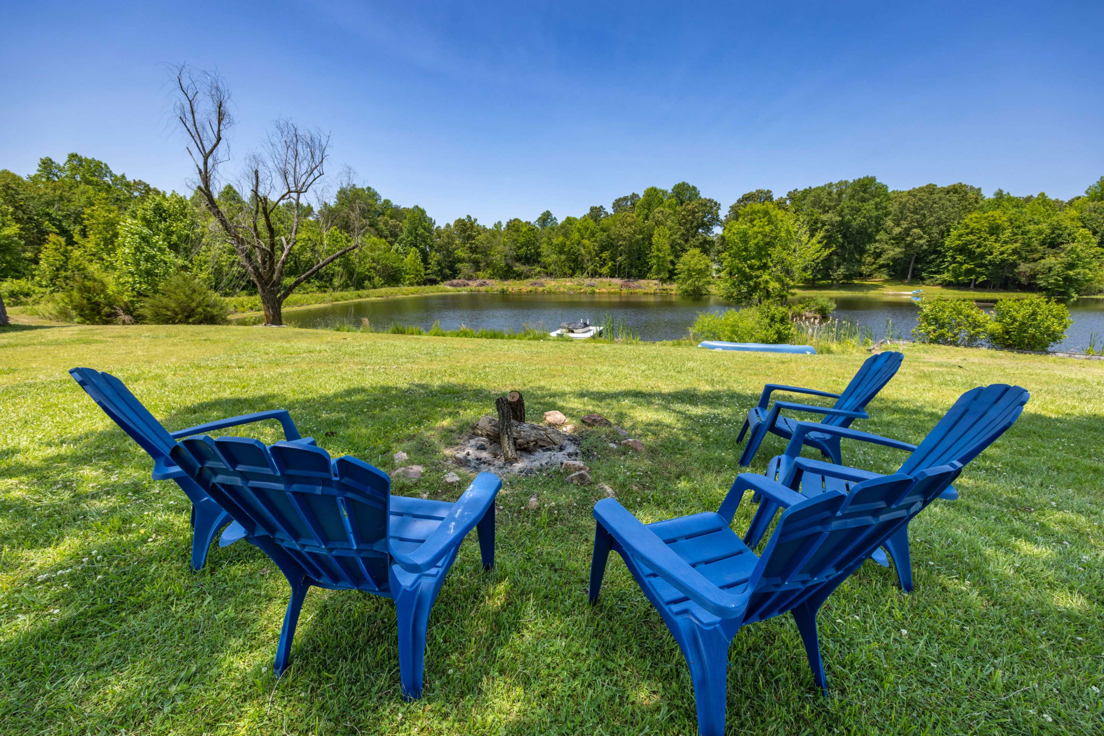 A circle of blue Adirondack chairs surrounds a small fire pit near a pond, with trees and greenery in the background.