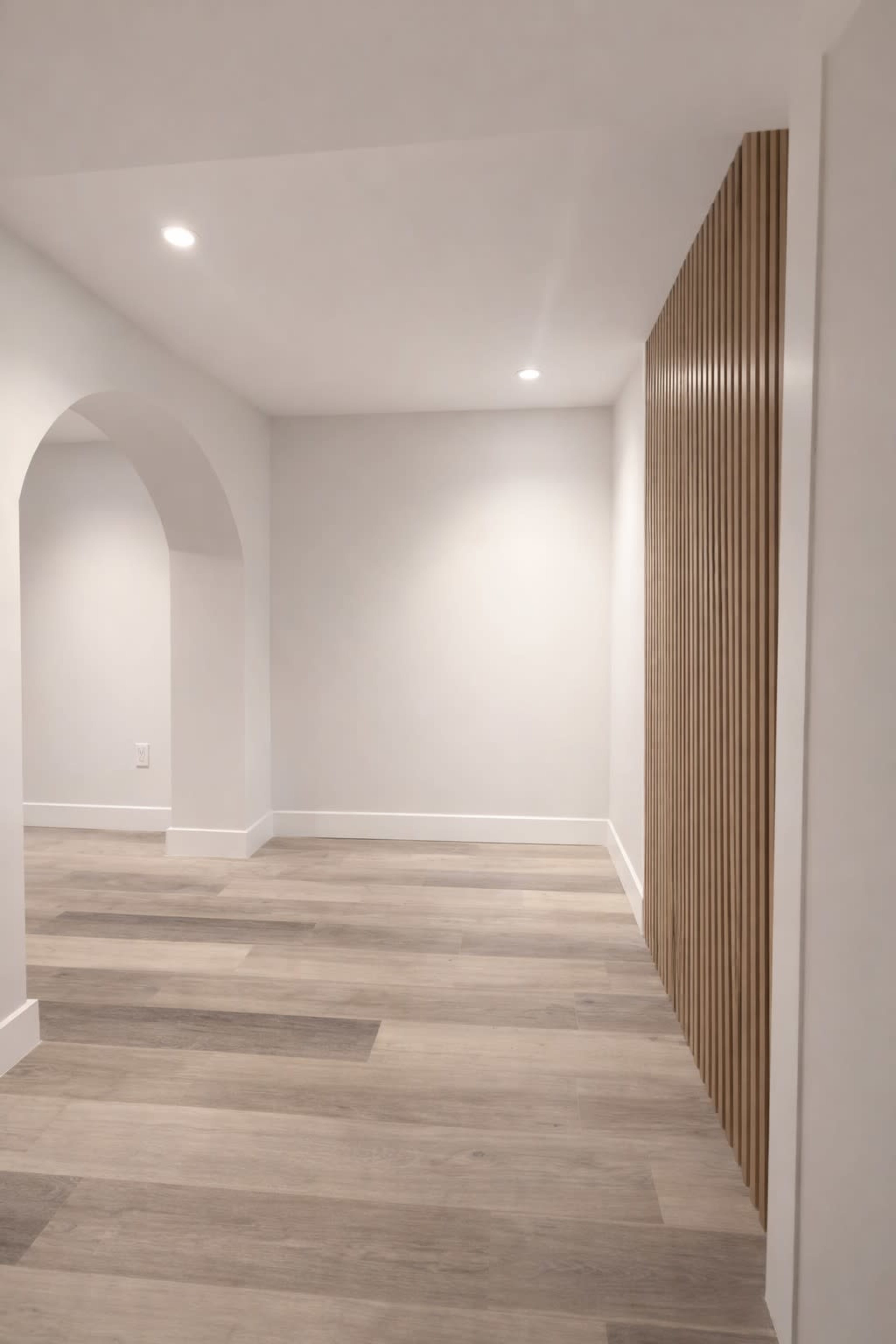 The image shows a spacious room with light wooden flooring, white walls, and an archway leading to another area, complemented by a vertical wooden slat feature on one side.